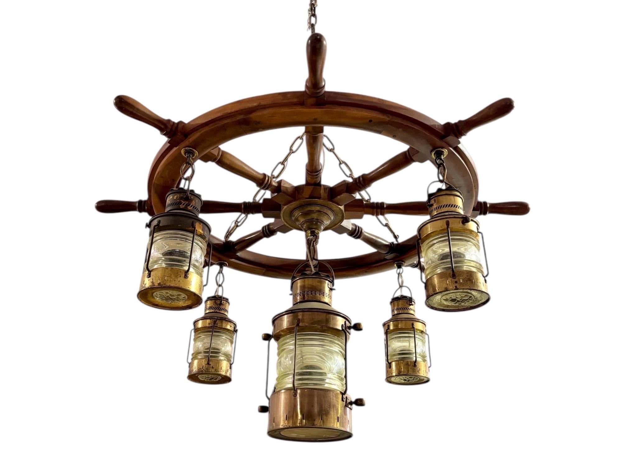 A mid-twentieth century nautical chandelier, made with an authentic solid wooden ship's wheel and five copper and beveled glass anchor lanterns. Four smaller lanterns hang from alternate handles of the ship's wheel, and a larger lantern hangs from