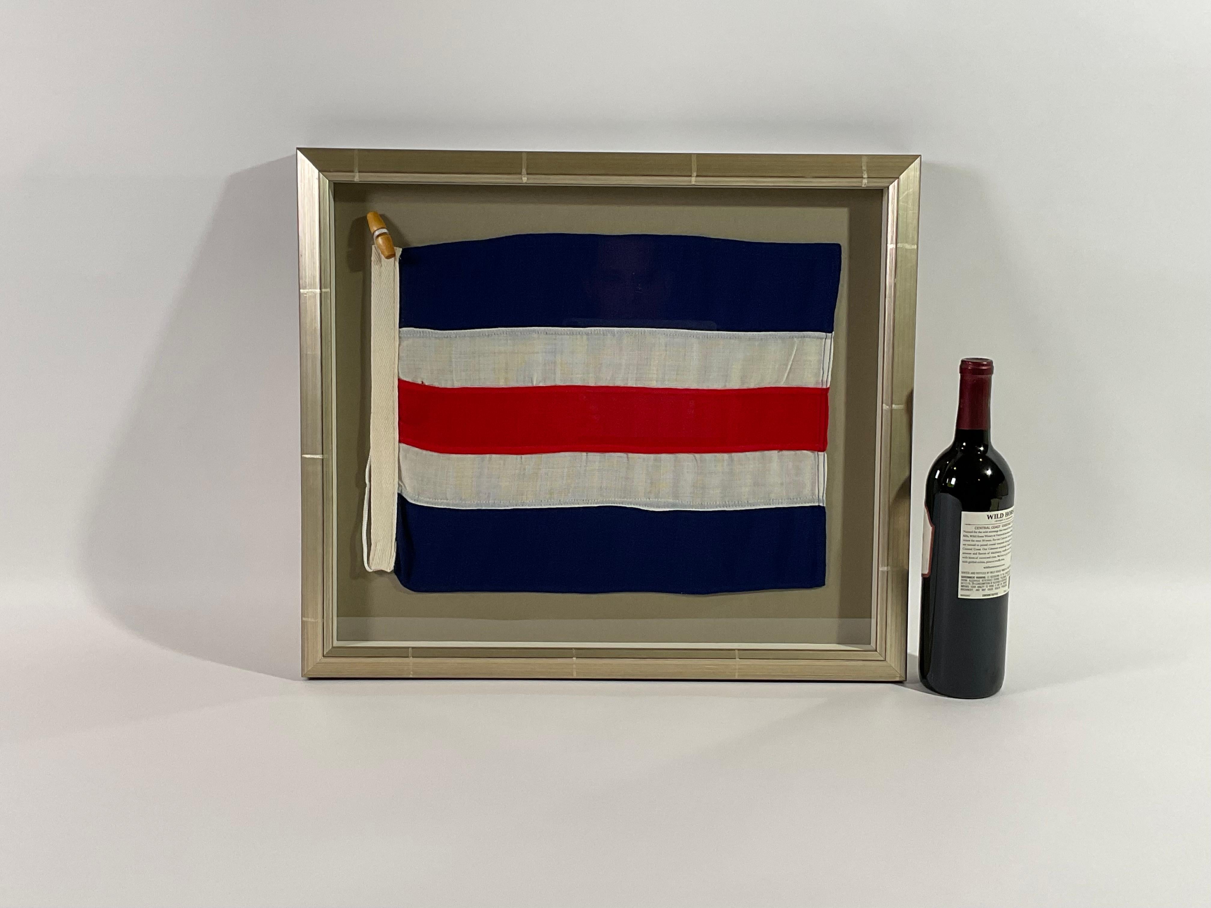 Framed maritime signal flag representing the letter “C” in the international code of signals.  This cotton signal pennant is made of individual stitched panels of red, white and blue with a strong canvas hoist band and original rope with wood
