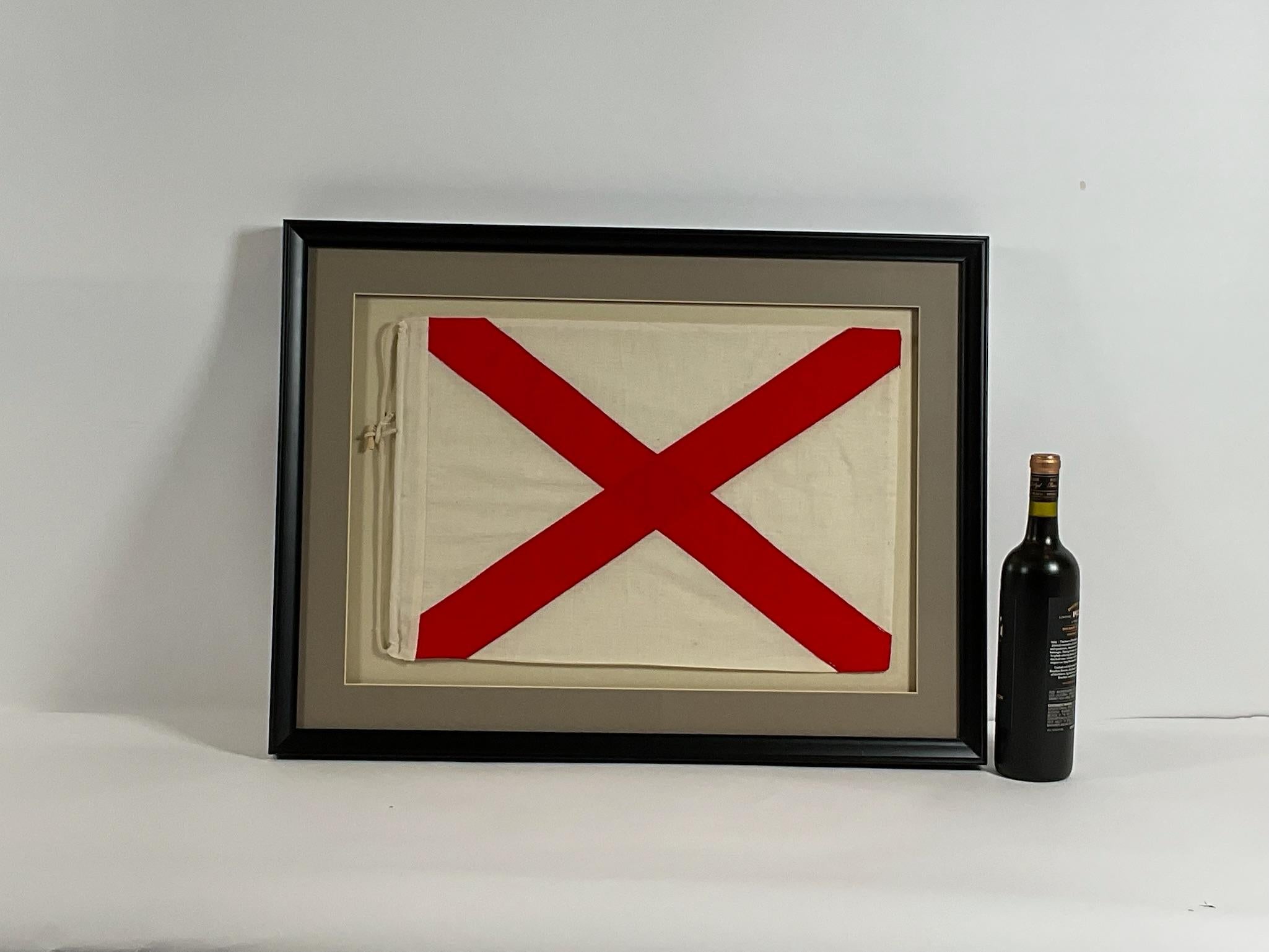 Maritime signal flag with rope and toggle fitted to a shadow box frame. White field with red X presenting the letter V for Victor. When raised, this flag symbols 