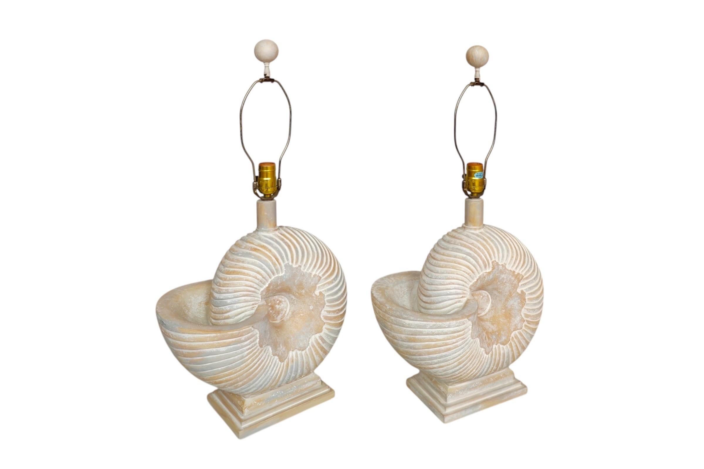 A pair of coastal style Nautilus shell table lamps. Made of plaster, detailed with ridged lines and finished in a sand color with gentle ash blue and tan tones. Harps are topped with matching ball finials. Each lamp measures 14