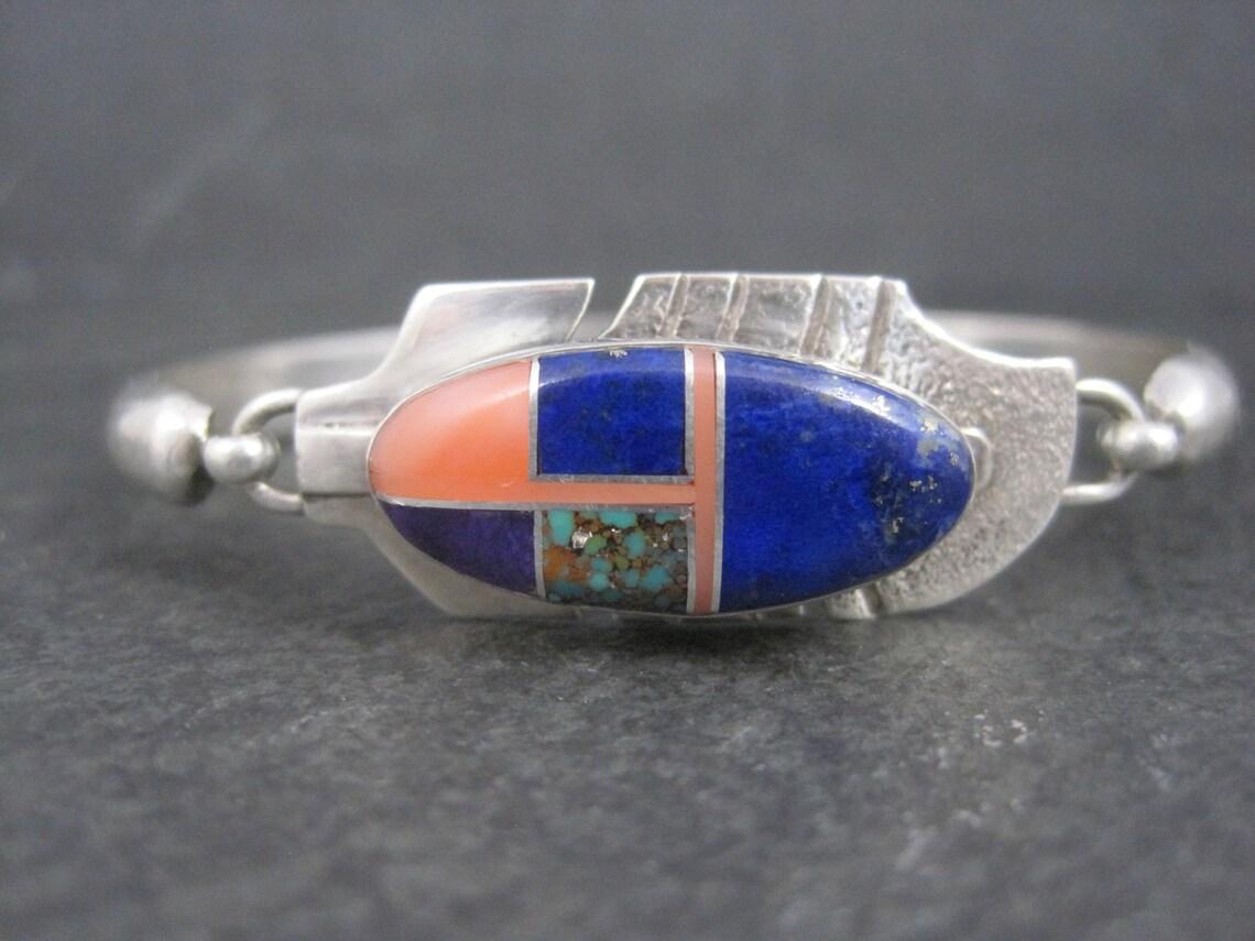 This gorgeous Navajo feather bracelet is sterling silver.

It features inlay in lapis lazuli, turquoise and pink shell.

The face of this bracelet measures 5/8 of an inch.
It has an inner circumference of 7 inches.
The hook is secure working