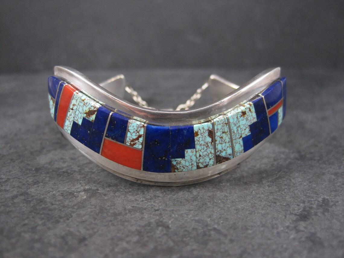 This gorgeous inlaid sweater cuff is sterling silver.
It features lapis lazuli, coral and turquoise inlay.

This bracelet measures 3/4 of an inch wide.
It has an inner circumference of 6 inches including the 1 inch gap.
It has a safety chain with