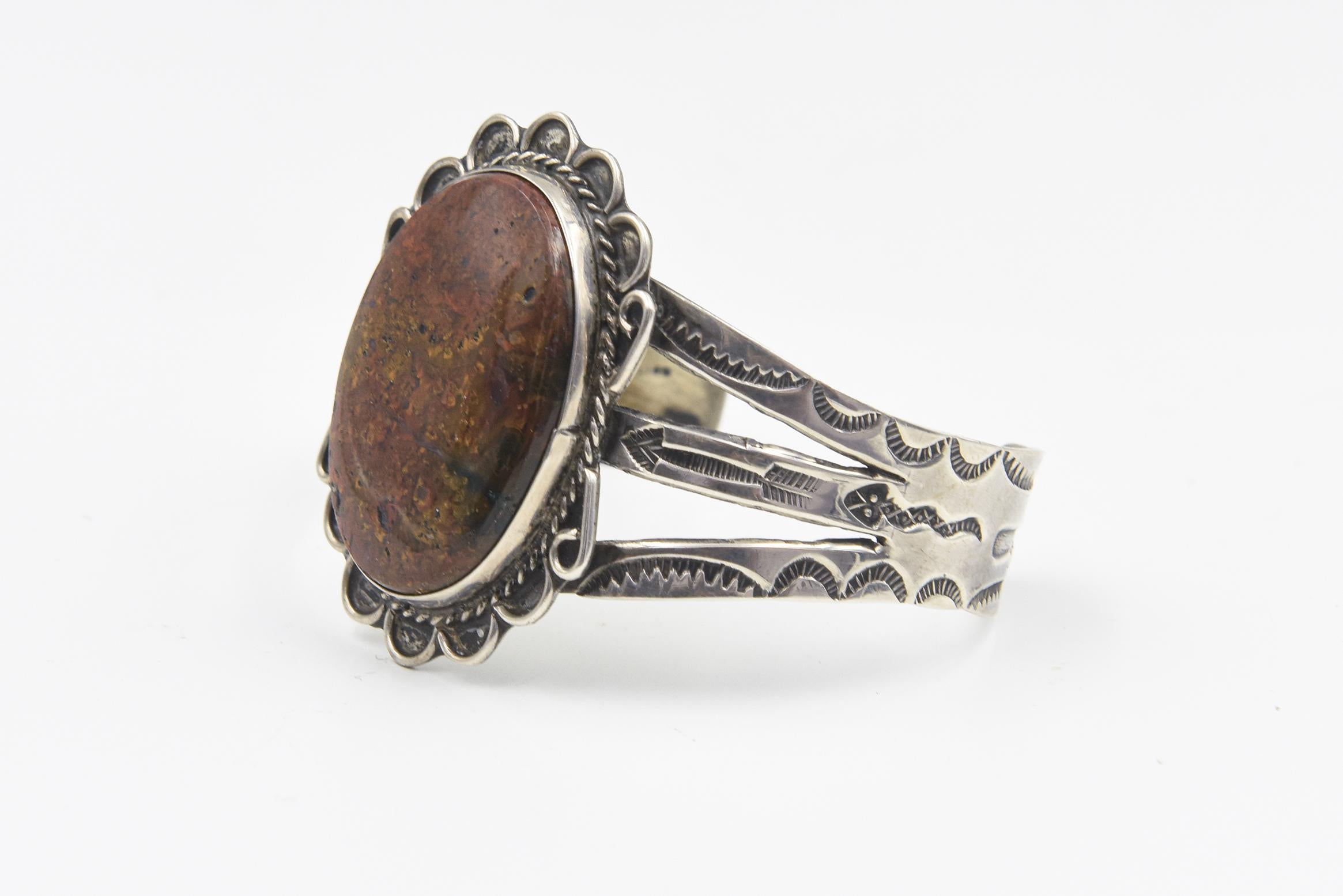 Bracciale in argento e calcedonio dei nativi americani. 
Il design del bracciale presenta un diaspro marrone ovale centrale in una cornice di girasoli con un bracciale a tre sezioni che include frecce, serpenti e cactus incisi.
L'area della pietra e