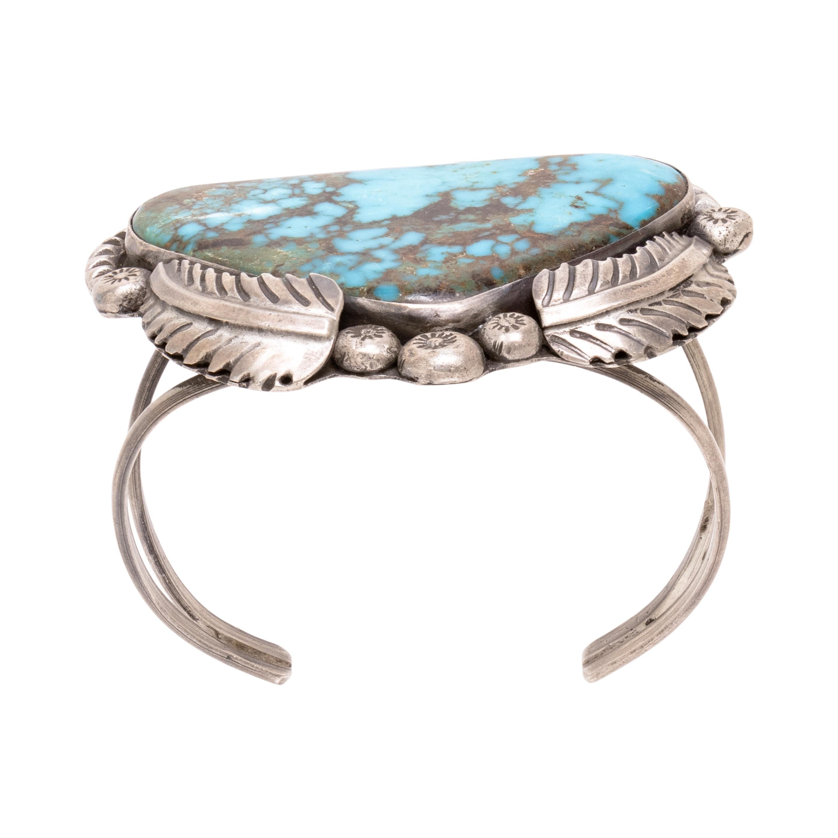 Pulsera navajo con una sola piedra turquesa, rodeada de hojas de plata y cuentas estampadas. Firmado R Lovato. La turquesa está tallada en forma de triángulo redondeado, con brillantes  Coloración azul con matriz marrón y dorada. La turquesa procede