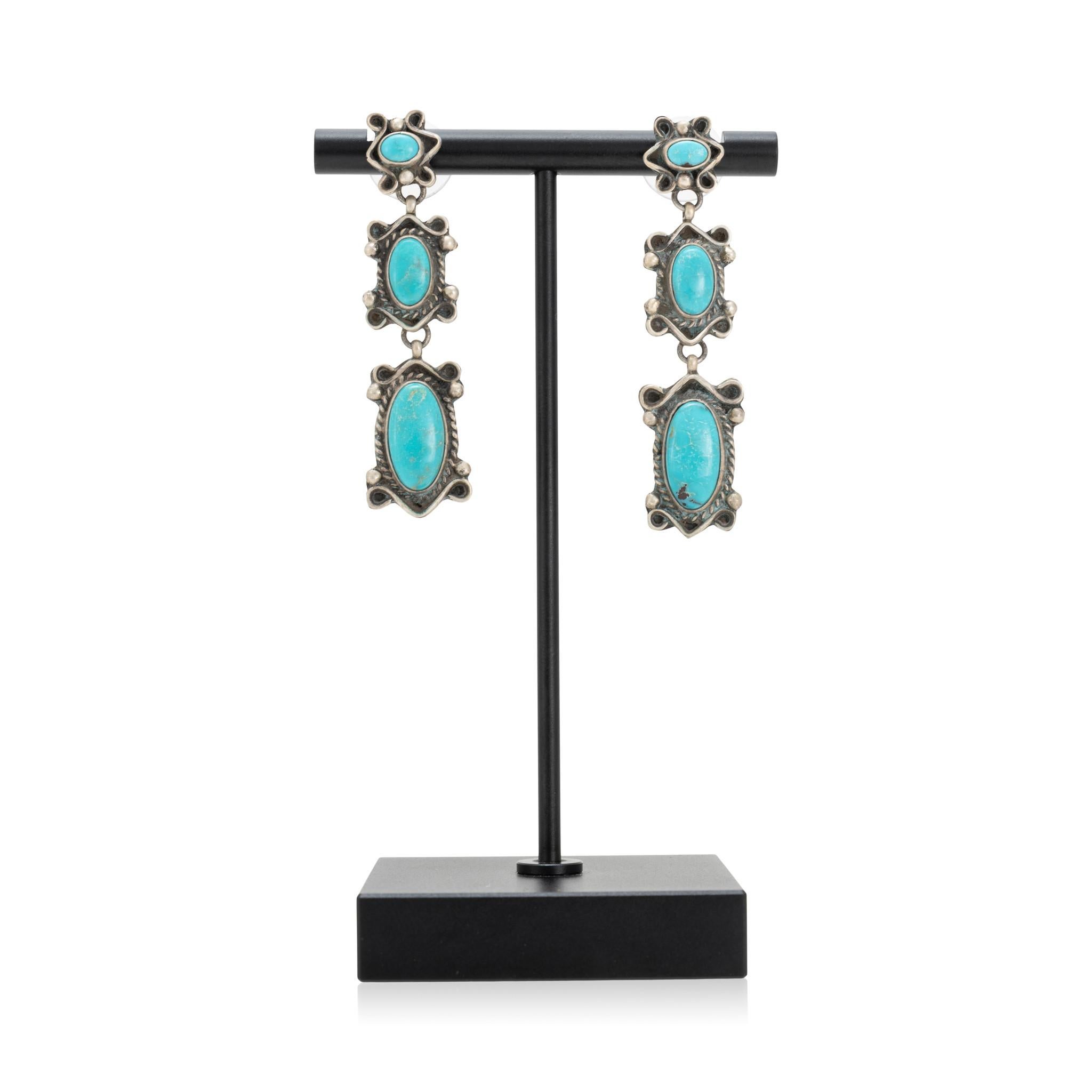 Orecchini pendenti in turchese Navajo Stormy Mountain e argento sterling. Tre pietre graduate con una colorazione blu brillante e una matrice bianca e marrone minima. Ciascuna pietra è incastonata in un castone liscio, circondato da una pergamena