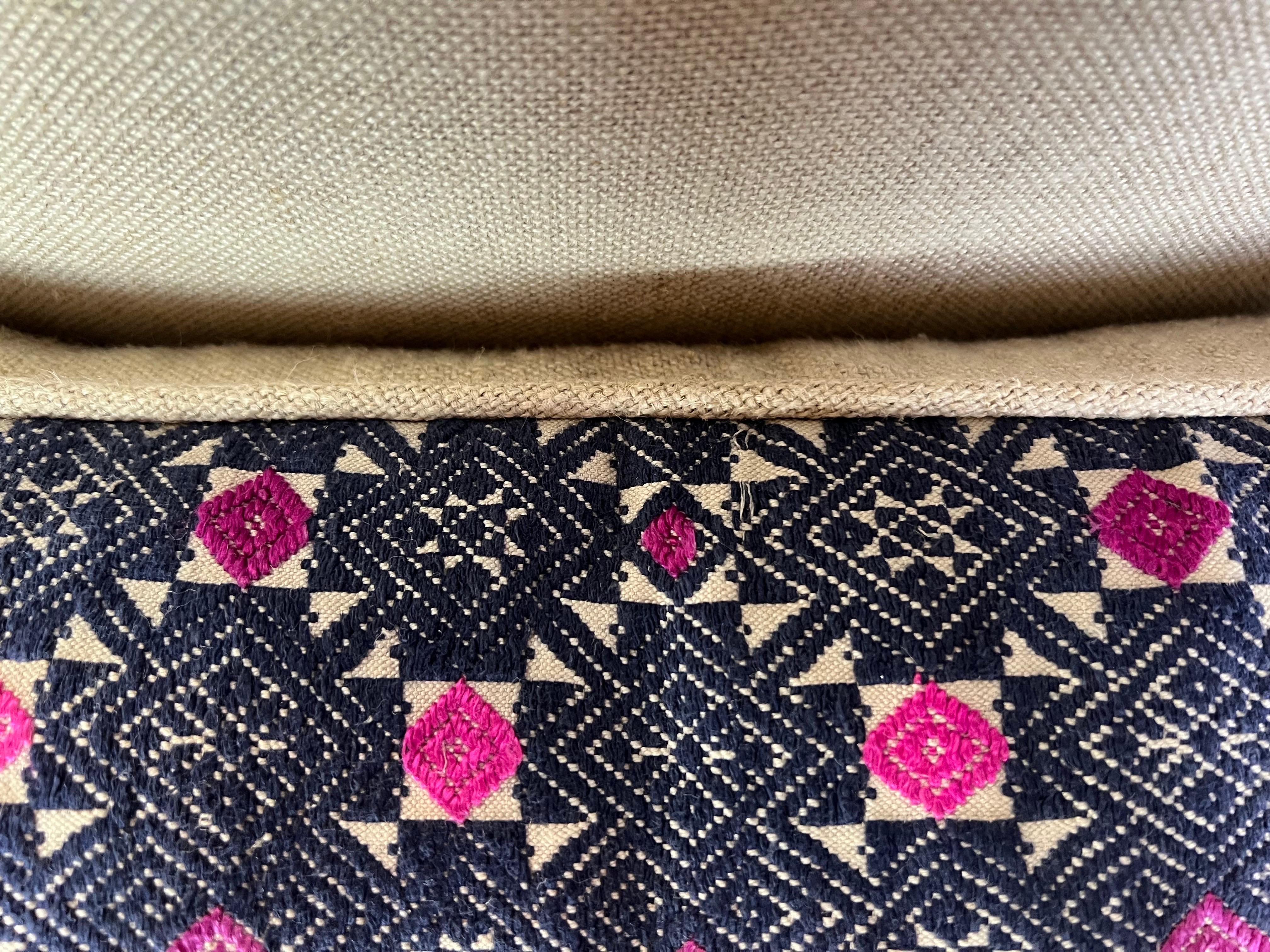 A decorative hand woven pillow with a geometric pattern.  The primary colors in the pattern are navy & pink, with the pink creating diamond-shaped highlights within the overall design.  There is a washed neutral linen back with a self cord detail. 