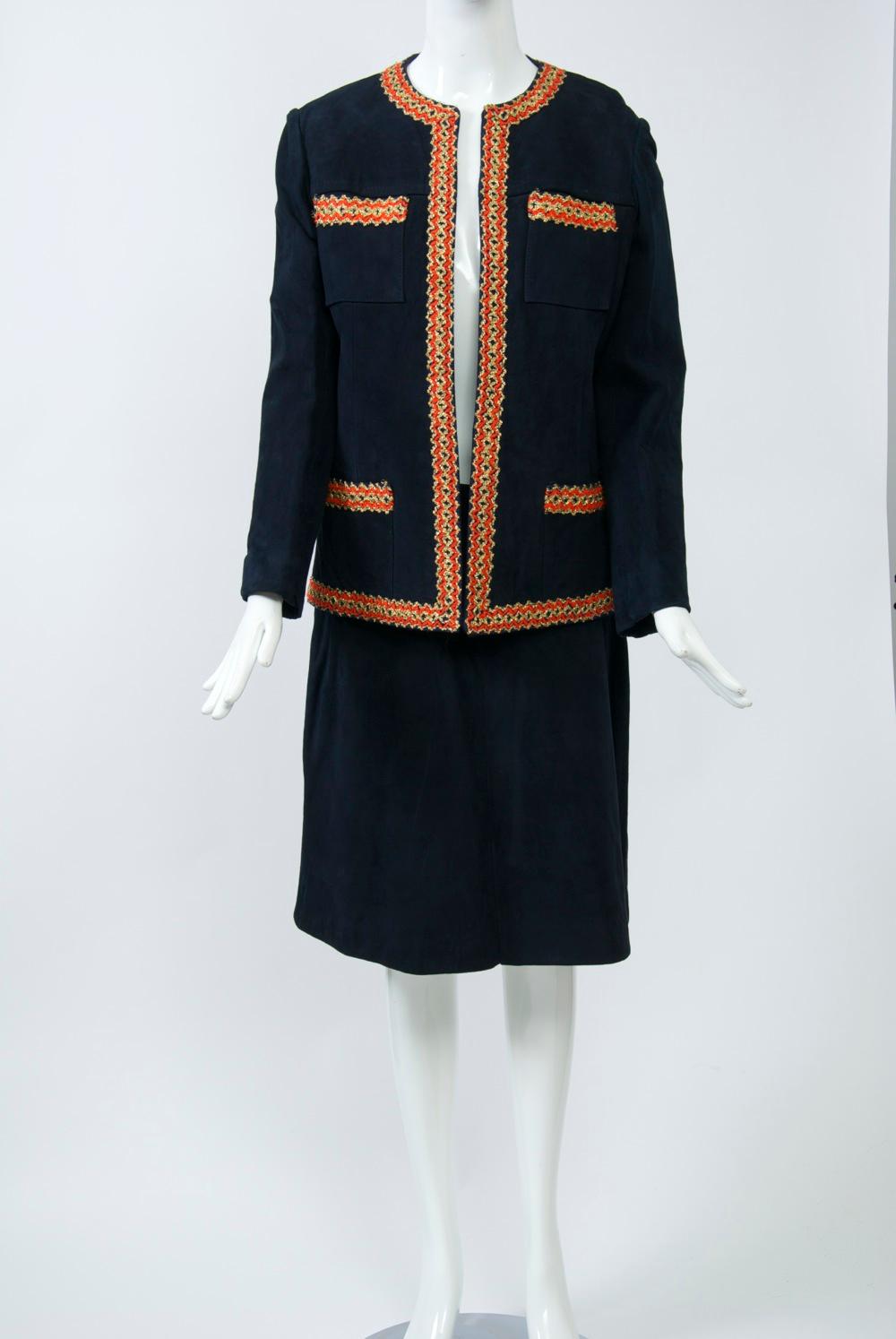 1960s navy suede suit in the style of a classic Chanel, it features a red and gold zigzag braid around the edges of the jacket and the top of the bodice pockets. The slim skirt rests on the waistline and has a slight A shape and back zipper. Jacket