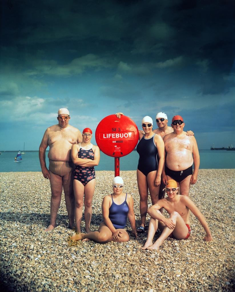 Channel Swimmers by Neal Slavin depicts an eclectic group posed by a red life buoy on a pebble beach. The group is dressed in swimming attire, all with varying colors and styles of goggles, swim caps and swimsuits. They stand in front of the deep