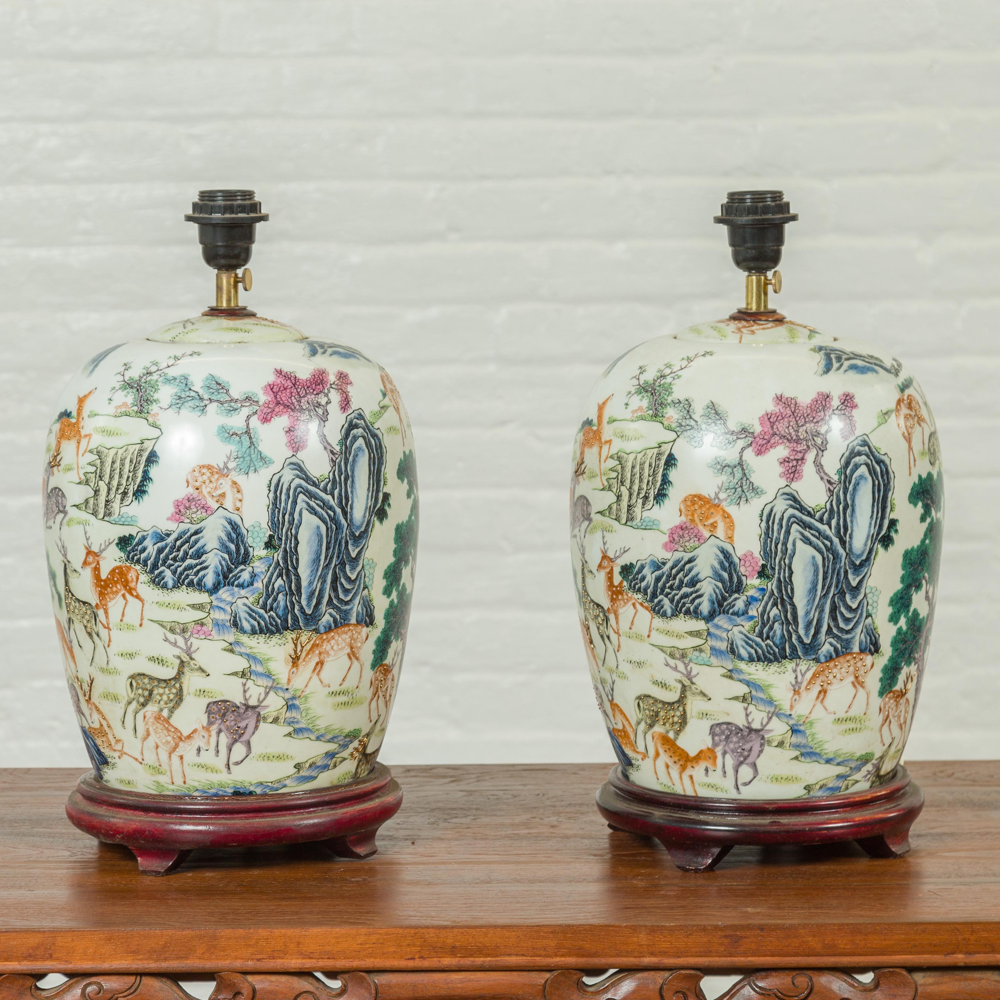 A near pair of Chinese deer in mountains table lamps from the 20th century, on wooden bases. Our eyes are immediately drawn to the delicate depiction of elegant deer and stags evolving inside a mountainous landscape highlighted with colorful trees.