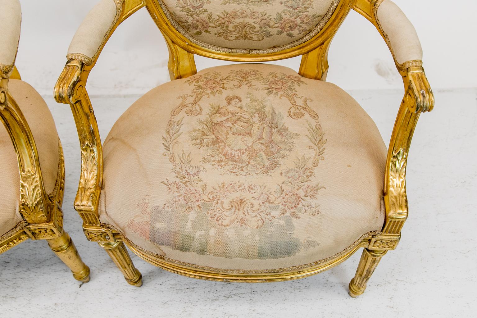 The fabric on this near pair of French gilt armchairs depicts courting scenes. There is significant wear and staining to the fabric and wear to the gilding in some areas. The oval backs have carved molded shapes and splayed arms have carved acanthus