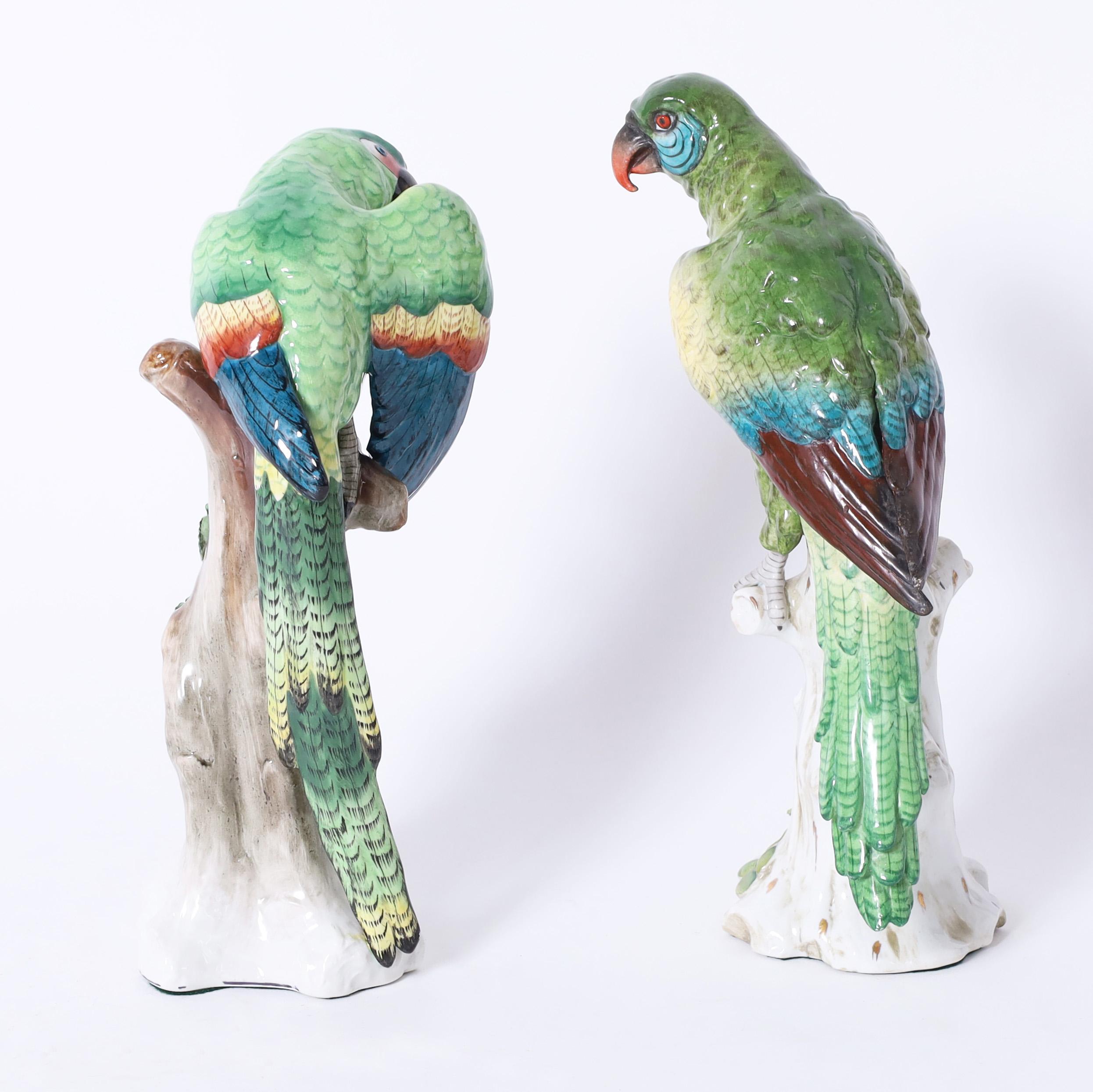 Standout pair of vintage life size porcelain green parrots both hand decorated in tropical colors, perched on tree trunks and signed on the bottoms.