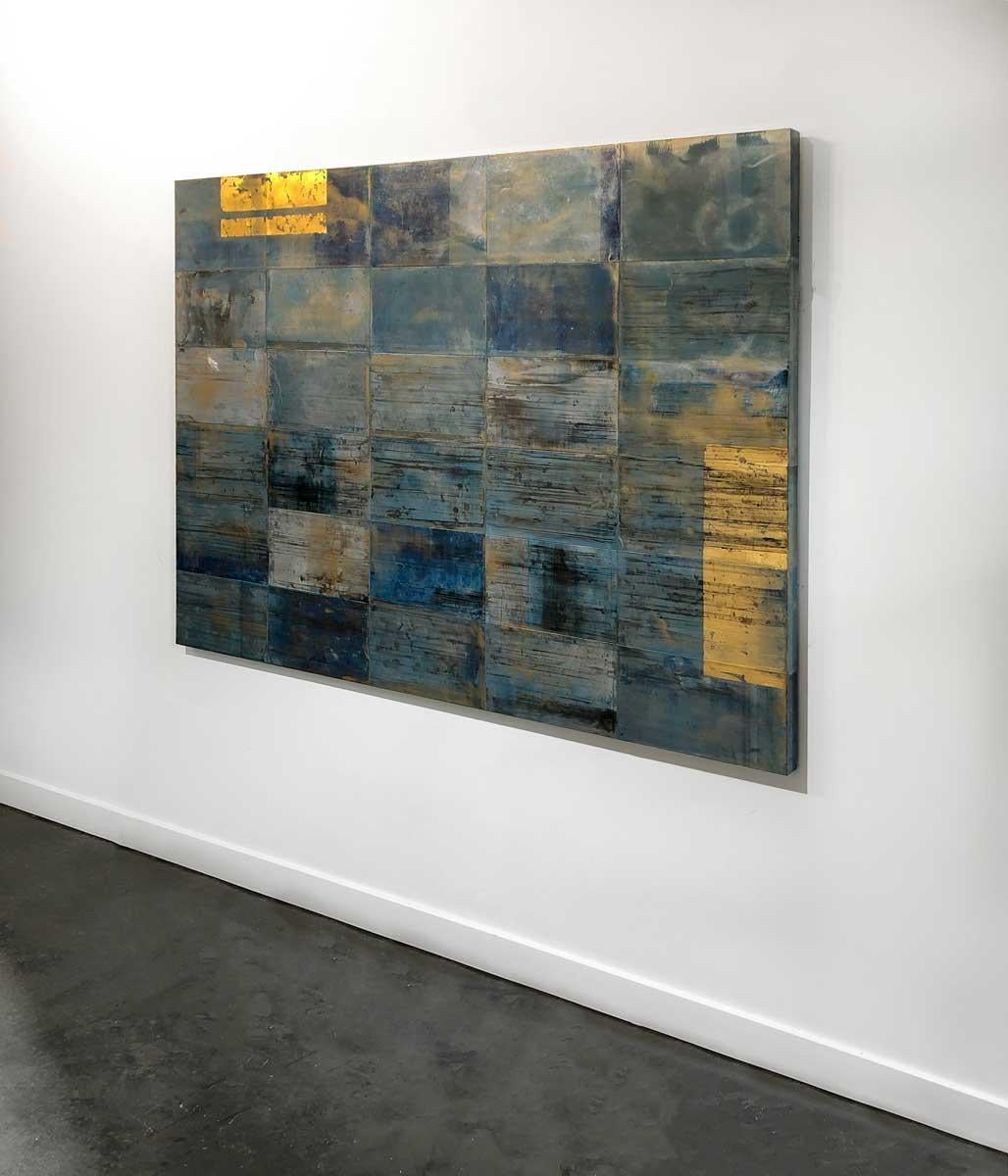 This large contemporary abstract painting features tiled blue and grey rectangles. They are textured with black and warm orange tones. Two bright metallic gold rectangles are layered on two edges of the piece. It can be hung vertically or