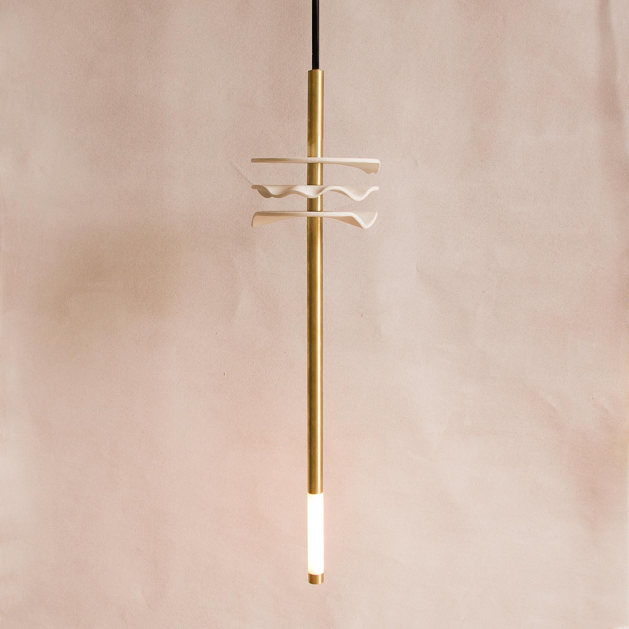 This dramatic pendant light features three organically shaped hand-formed white porcelain disks, each with an entirely unique wave pattern. The porcelain disks are pierced by a slender unfinished brass rod with integrated LED, and hands from a black