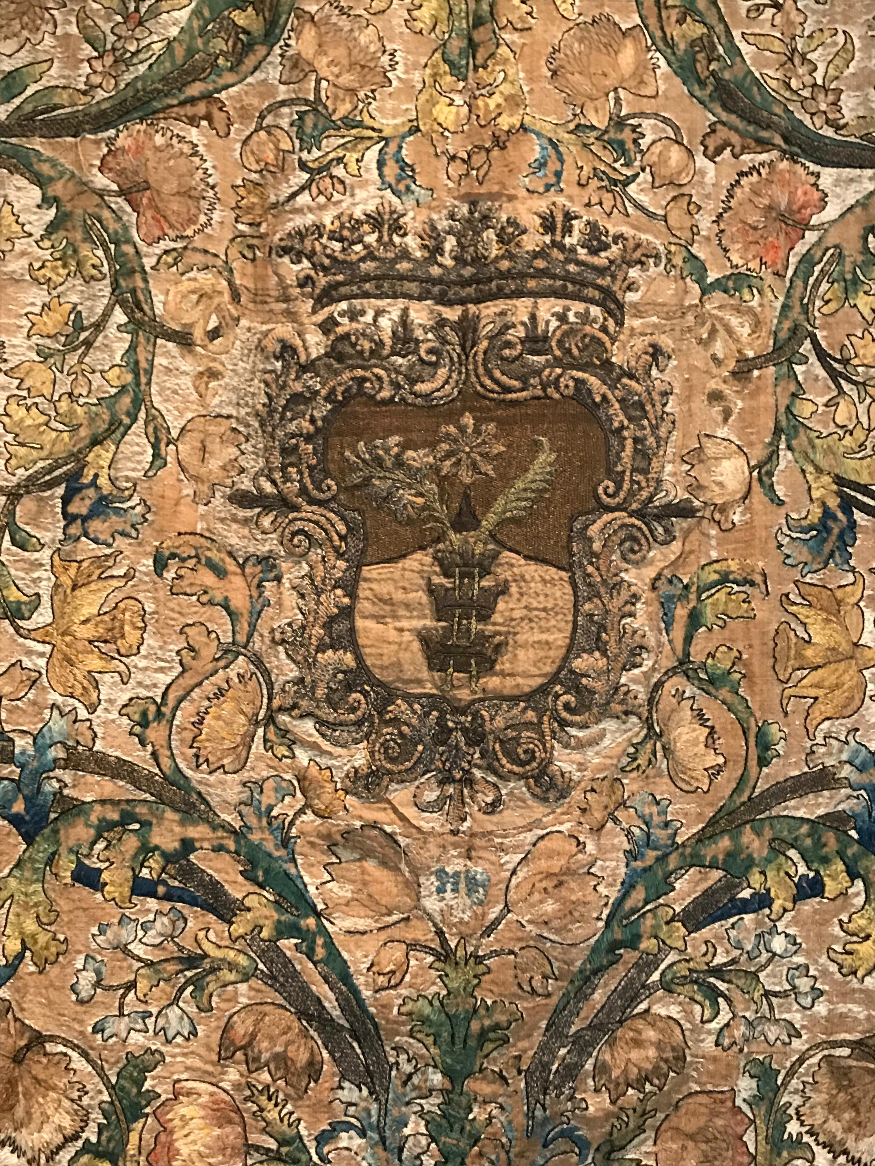Beautiful needlework tapestry with intricate floral and vine patterns and a shield in the center. Shades of yellow, green, brown and blue with a brown border.