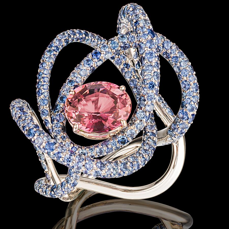 The Kephi Ring by Neha Dani reflects passion in a 4 Carat Reddish Pink Oval Natural (no heat) Sapphire, which is surrounded by 6.5 Carat Round Full Cut Blue Sapphires. Set in 18K White Gold, the ring exudes elegant brilliance.