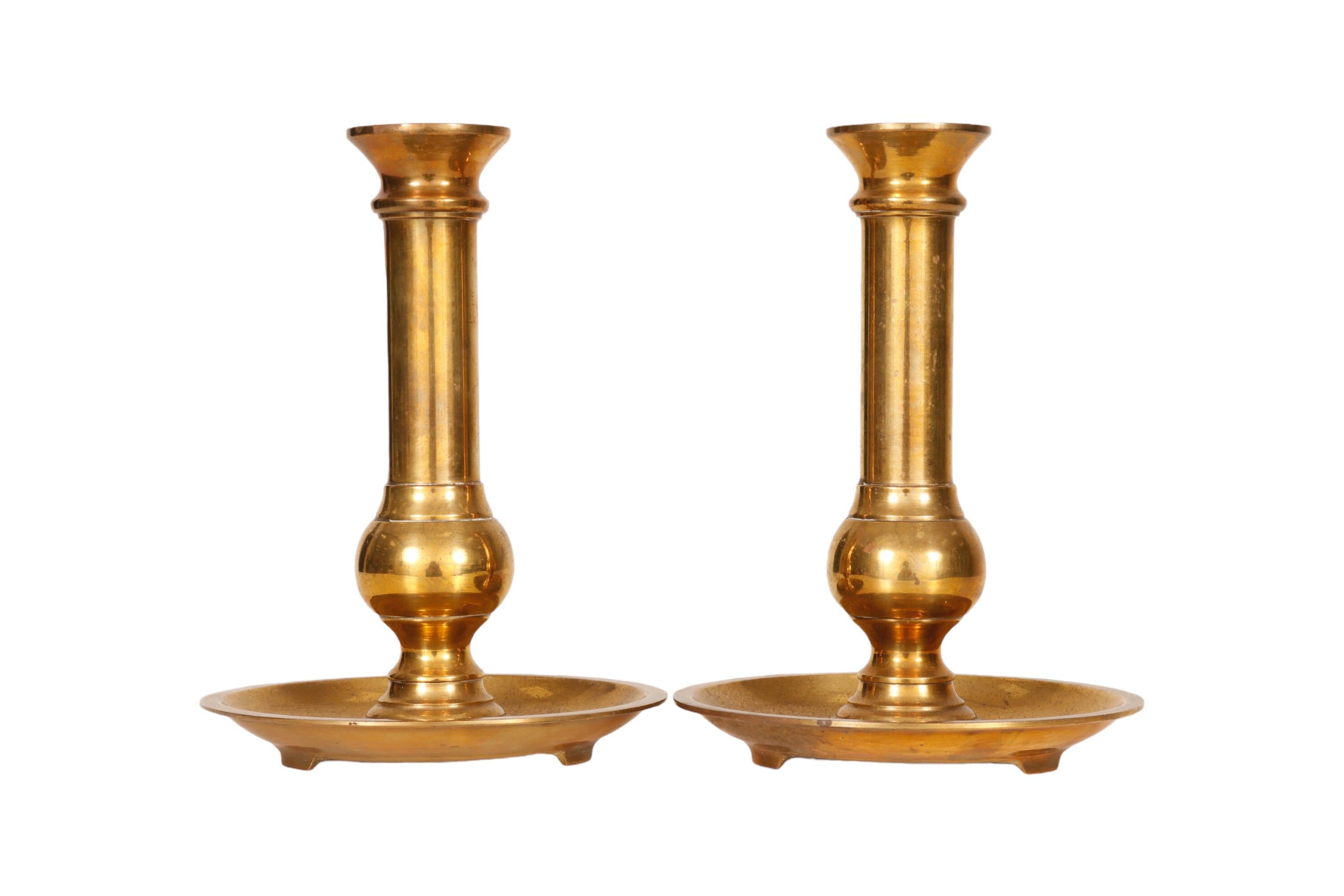 A pair of traditional brass candlestick holders made by Neiman Marcus. Simple columns with ball shaped turned bases sit in wide drip pans, each with three feet. Maker's label can be seen underneath. Dimensions per candlestick.