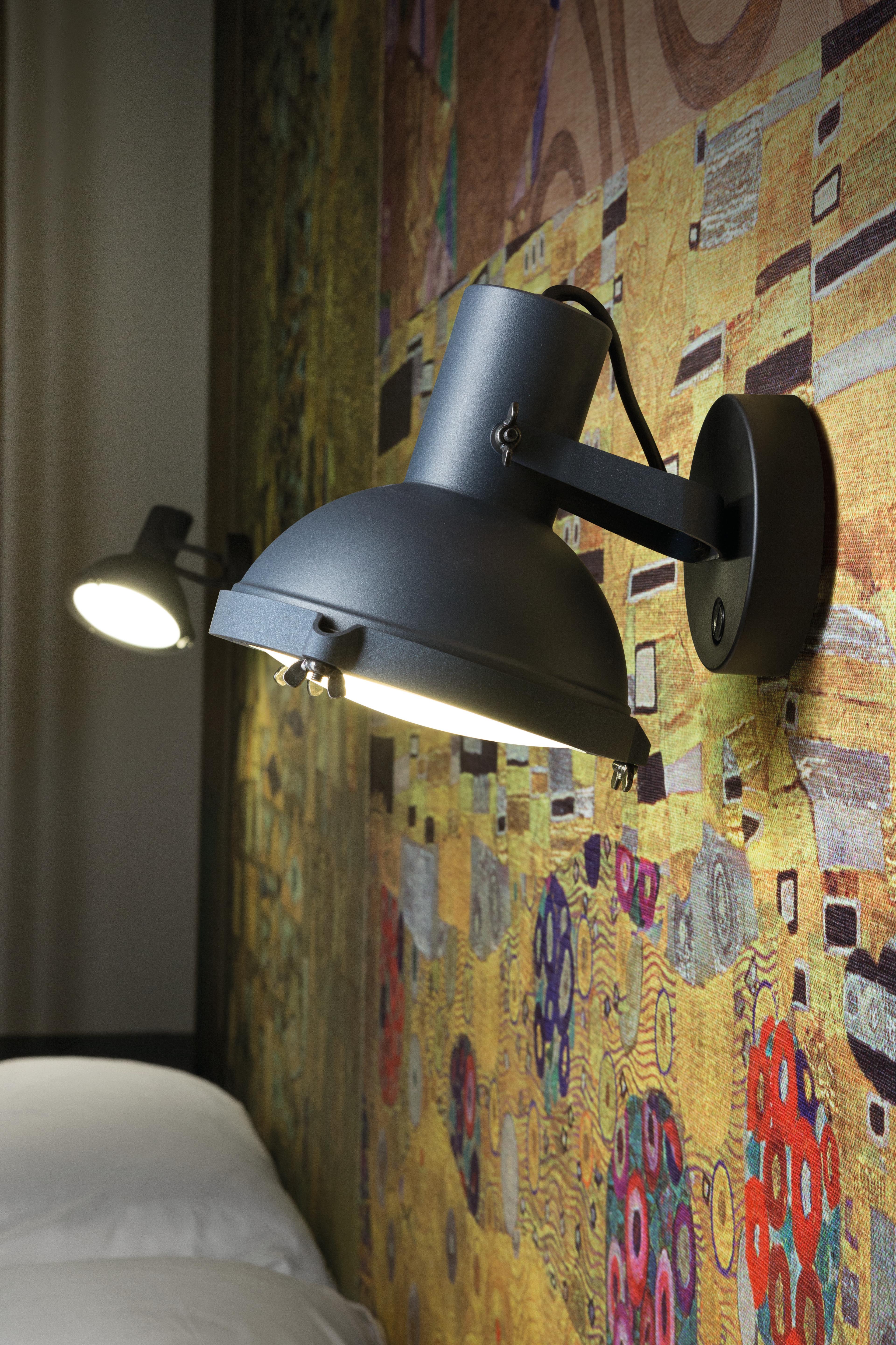 Lámpara LED de sobremesa de aluminio extruido con soporte en forma de T de polímero relleno de fibras de vidrio. Compuesto por un brazo saliente pintado de negro mate donde se encuentra la fuente de luz y un contrapeso vertical de latón. La lámpara,
