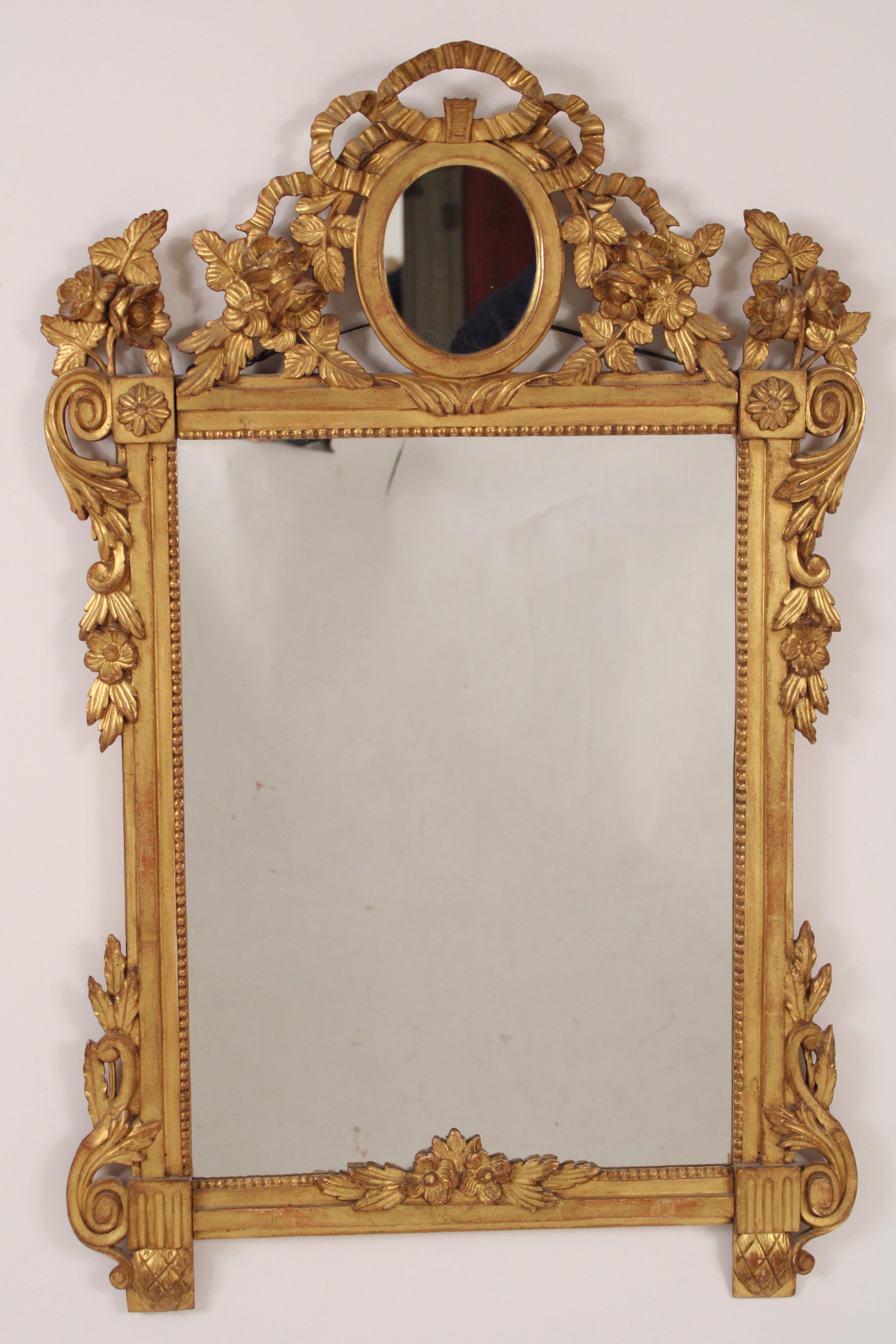 Antique Louis XVI style gilt wood (gold leaf) mirror, circa 1900. The mirror's pediment with ribbon, leaf and foliate wood carvings surrounding an oval mirror. The mirror's stiles with leaf and foliate gilt wood carvings.