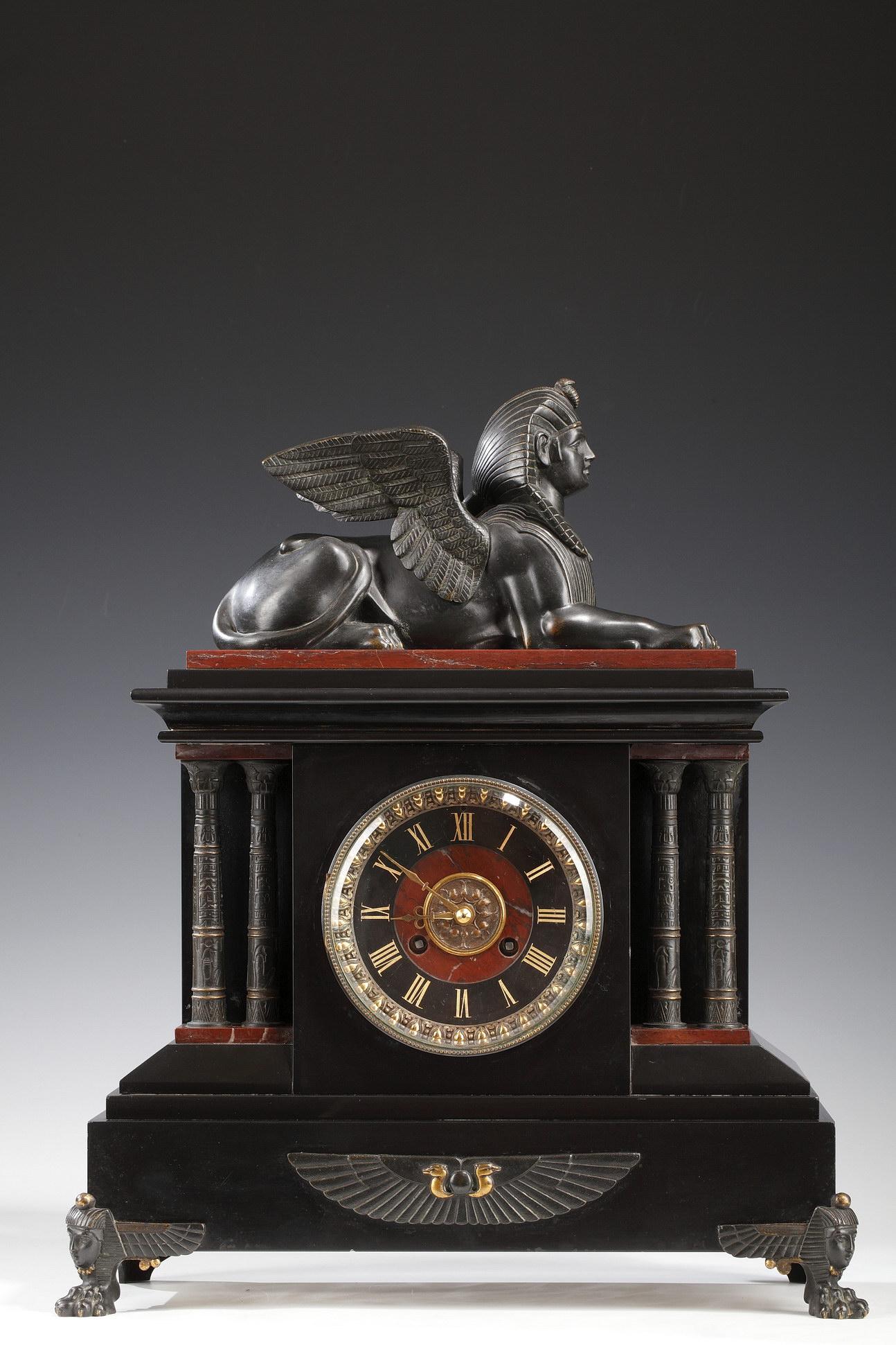 Reloj de mármol negro y rojo con forma de templo egipcio atribuido a G. Servant, flanqueado por columnas dobles inscritas con jeroglíficos y ornamentadas con un uraeus alado, todo ello realizado en bronce patinado a dos aguas. El reloj está coronado