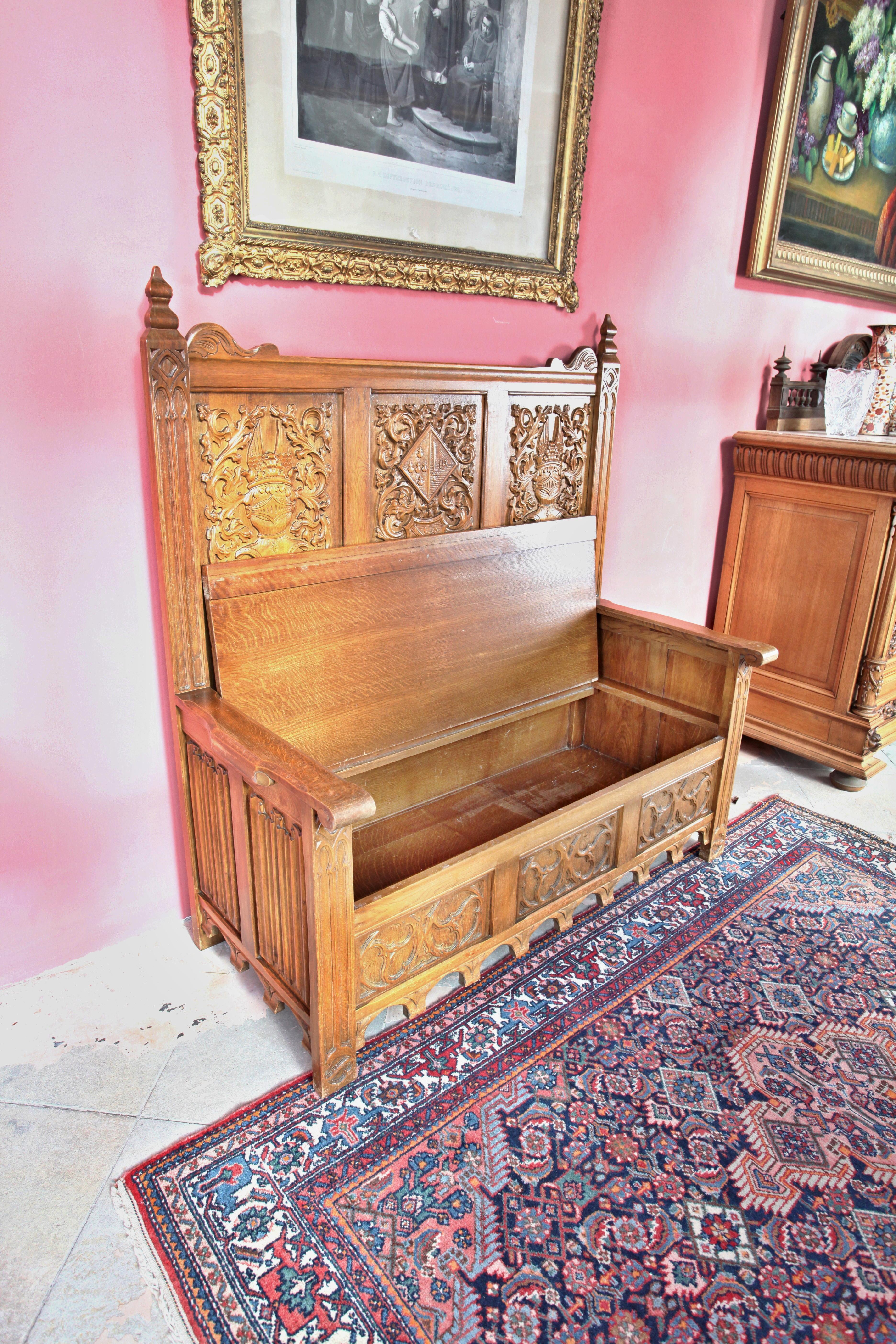 A solid oak bench richly decorated with hand-carved Gothic style. There is storage space inside the bench. There is an organ pattern on the sides and the frontal coffered panels are carved with a floral motif surrounding the coats of arms.
