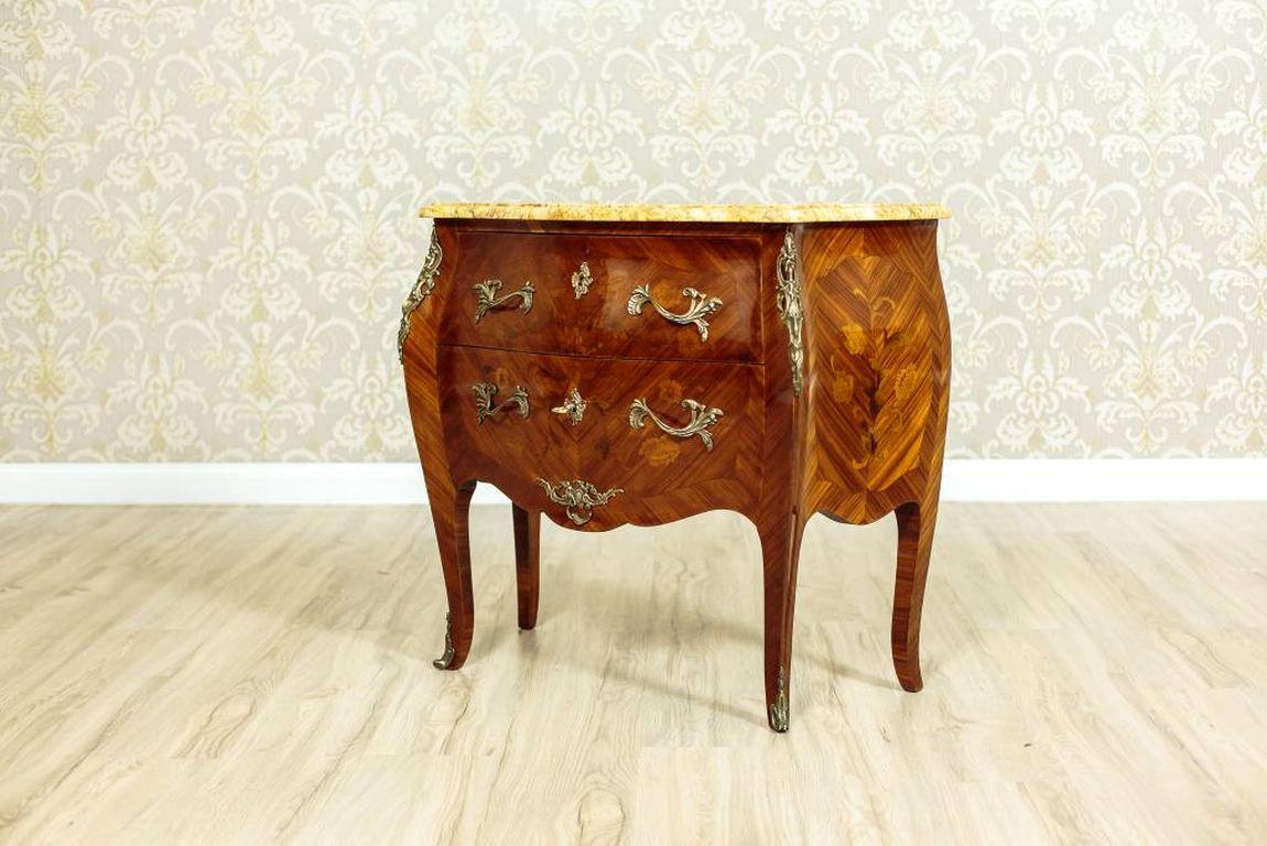 This dresser, circa 1930, resembles furniture in the Louis XV style.
This piece of furniture in the French type has a bulbous shape and a marble top, which is ornate with marqueteries and intarsias.
The handles of the drawers and the appliqués