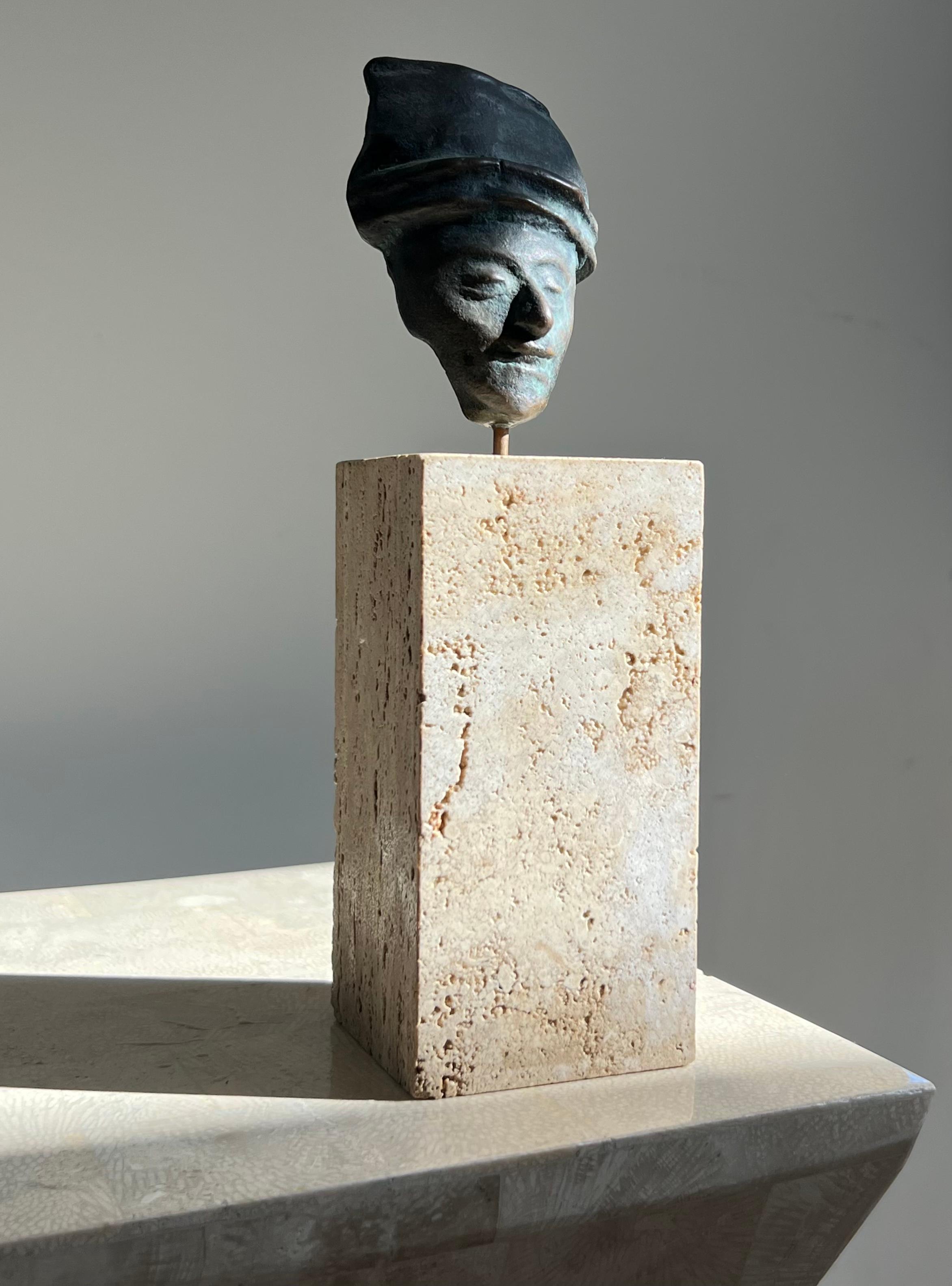 A Neo Sumerian style bronze sculpture of a face, mounted on a raw travertine plinth, 20th century. The felted bottom also shows a sticker from a now shuttered antique store in Santa Monica where the relic was once sold; beyond that, the piece is