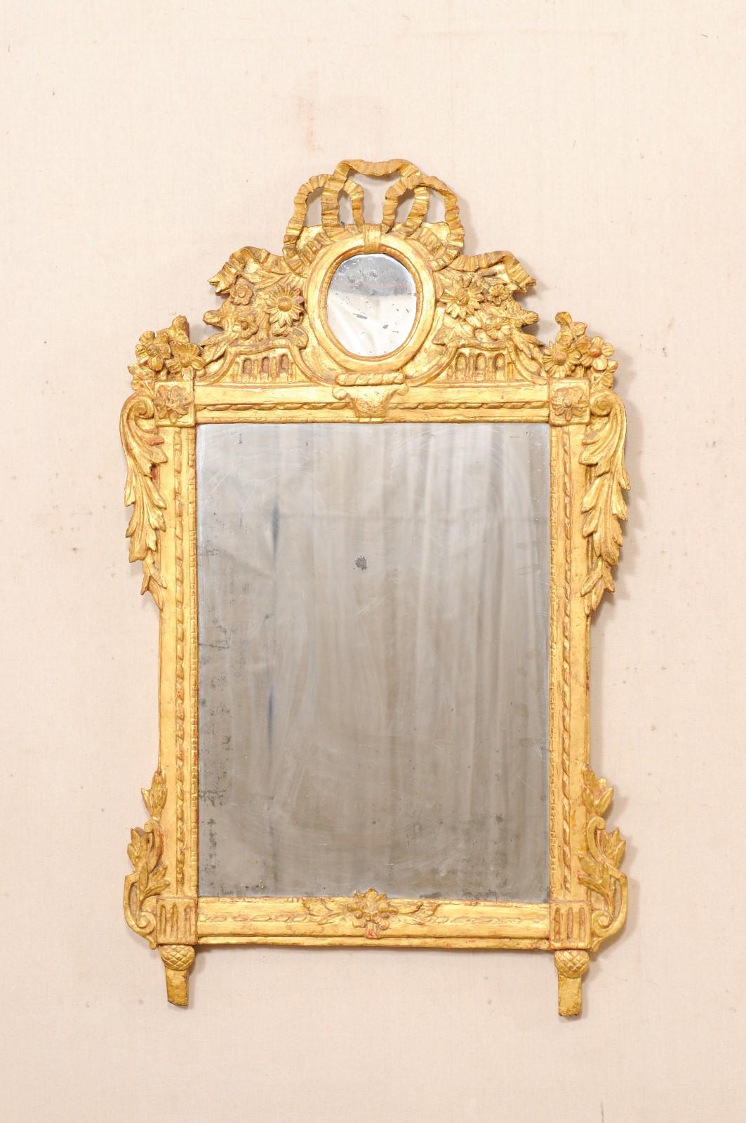 A French Neoclassical carved and gilt wood mirror from the 19th century. This antique mirror from France features a beautifully carved crest with a smaller, round-shaped mirror at center, with a bow-tie at top and cheerful flowers surrounding it at