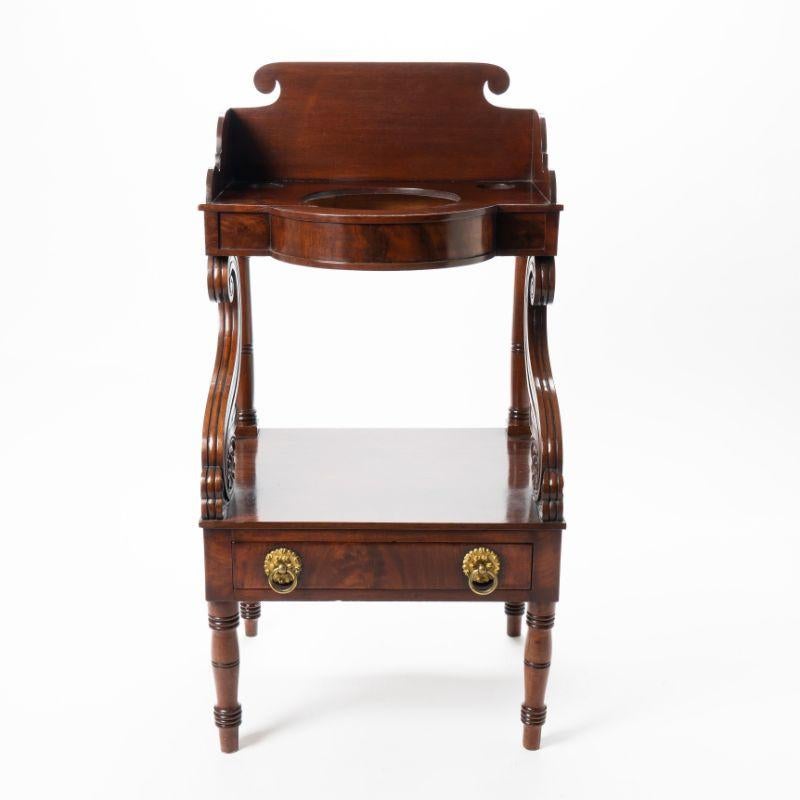 American Neoclassic mahogany wash stand. The stand has a bowed basin shelf on a conforming apron with scroll cut splash. The lower stretcher drawer is mounted with paired and stamped gilt brass drop ring pulls. The front of the upper shelf is