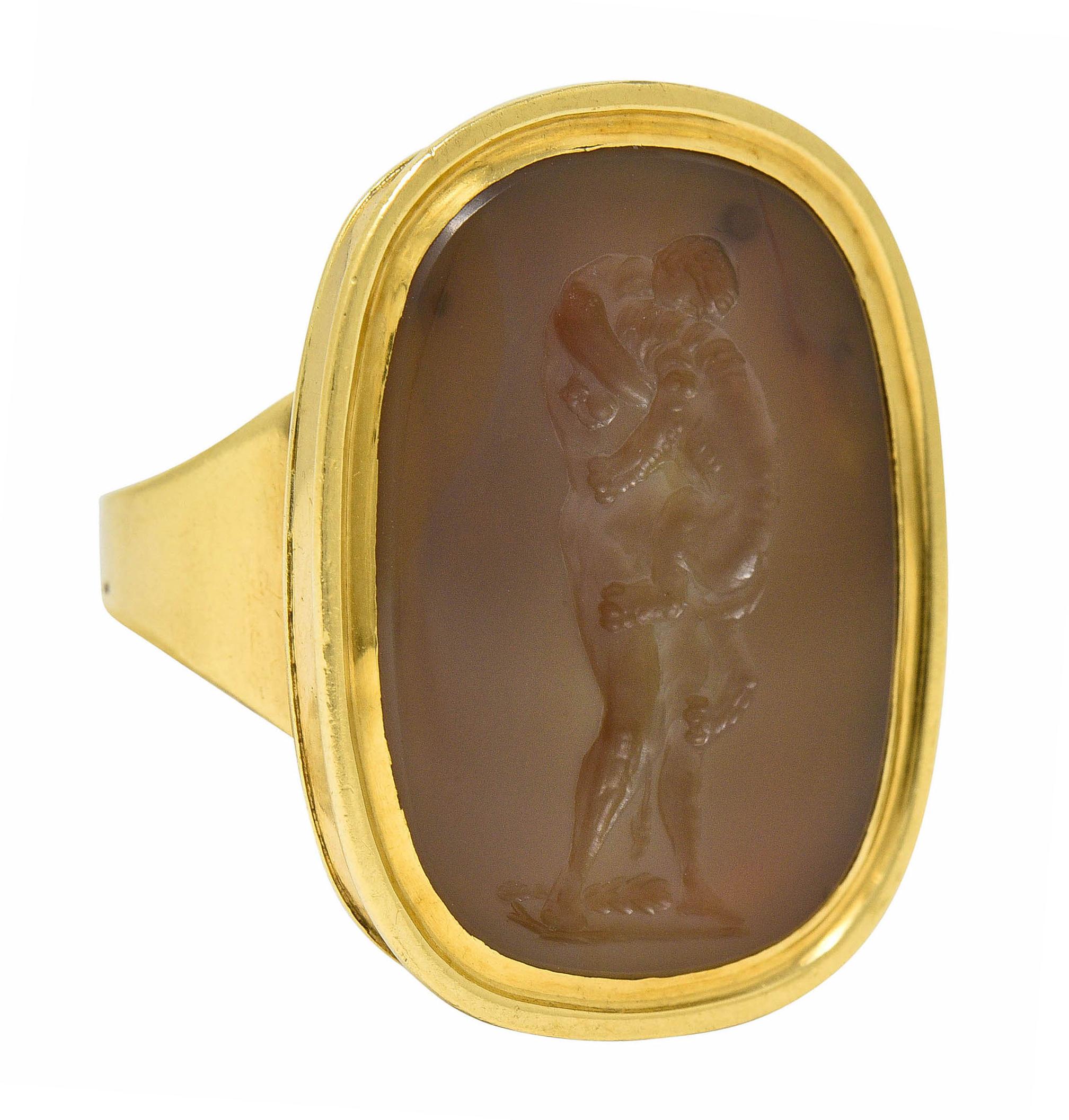 Centering an elongated cushion tablet of agate that measures 25.0 x 19.0 mm

Translucent taupe in color with very subtle mottling

Deeply engraved to depict Hercules grappling with the ferocious Nemean lion

Bezel set in a polished gold