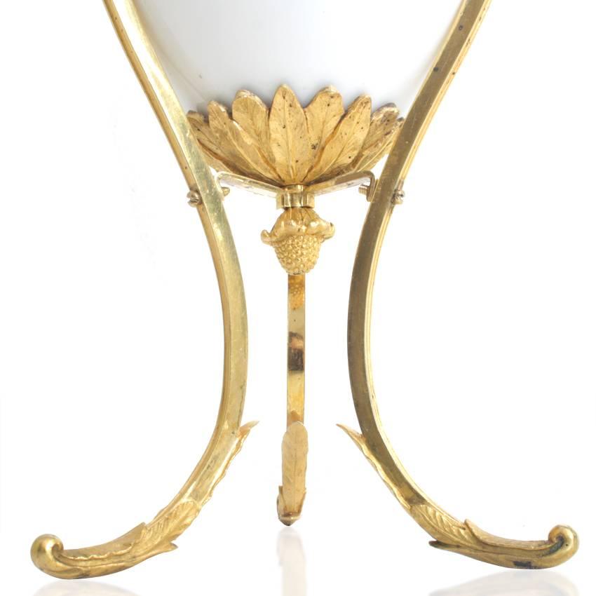 TThe glass vase is surrounded by a tripod, whose legs run out into leafy vines and whose handles are designed as goosenecks. The lower part is held together by three cross-connections, which terminate below a leaf frieze in which the vase body rests