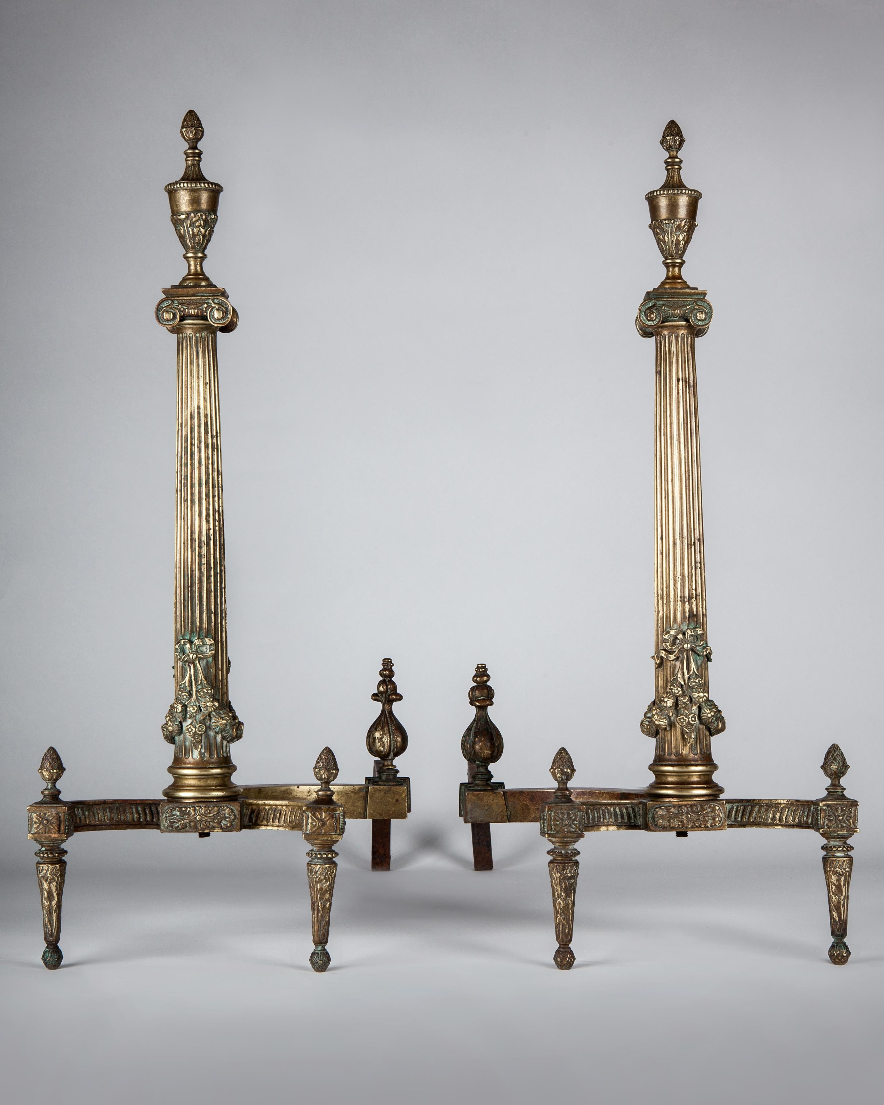 AFP0584
A pair of antique cast brass andirons having fluted tapered columns mounted with floral swags and ionic capitals, surmounted by urn finials. The column shafts rest on an inward curving set of legs and wrought iron supports clad in brass are