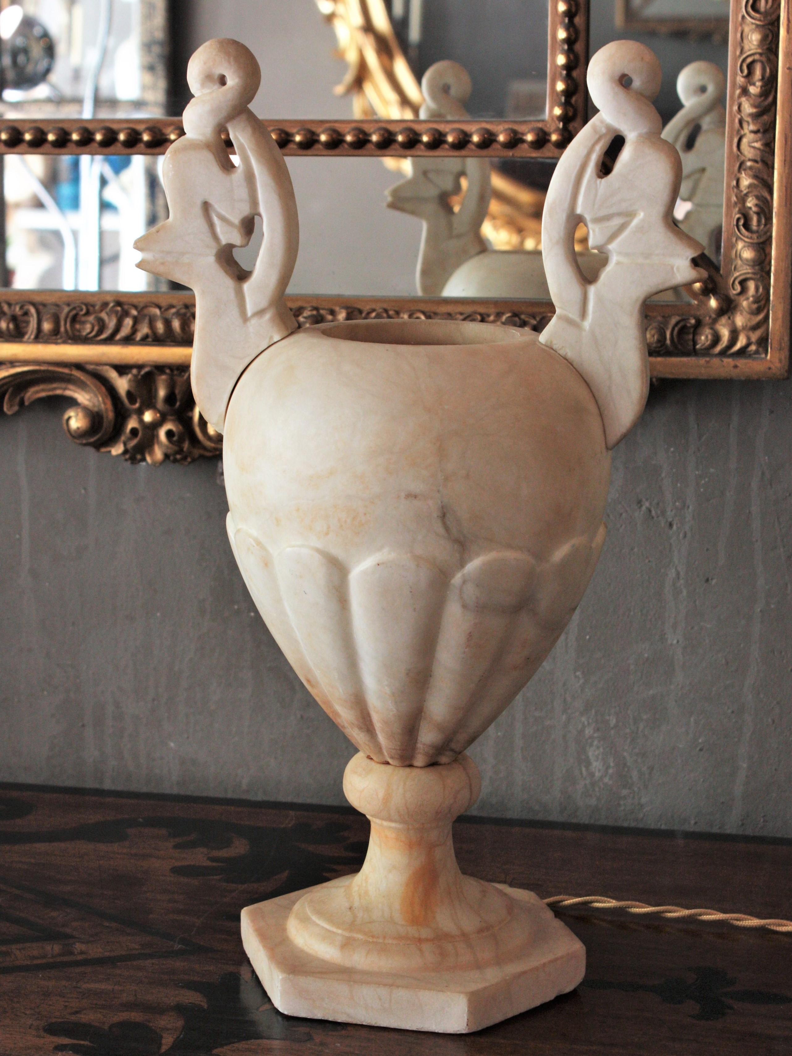 Sculptural carved alabaster Art Deco period urn table lamp with neoclassical design flanked by carved handles. Spain, 1930s.
It has carved decorations on the bottom part and hand carved handles adorning the top at both sides. 
Impressive