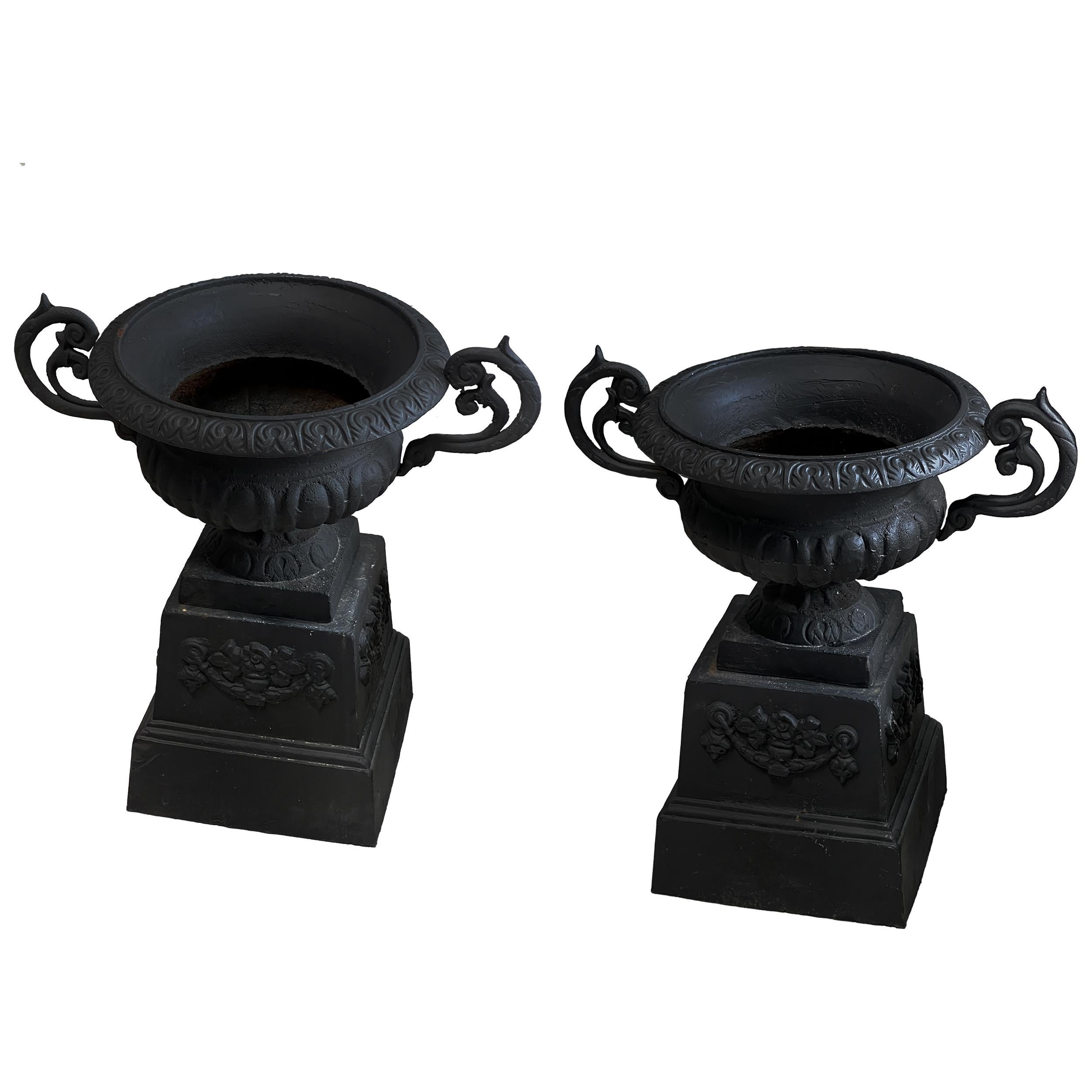 The Neoclassical Black Cast Iron Urns on Base/Plinth, are a captivating set of two garden urns / planters featuring c-scroll handles that exude timeless elegance. These urns bear a natural patina consistent with their antique age and usage, adding
