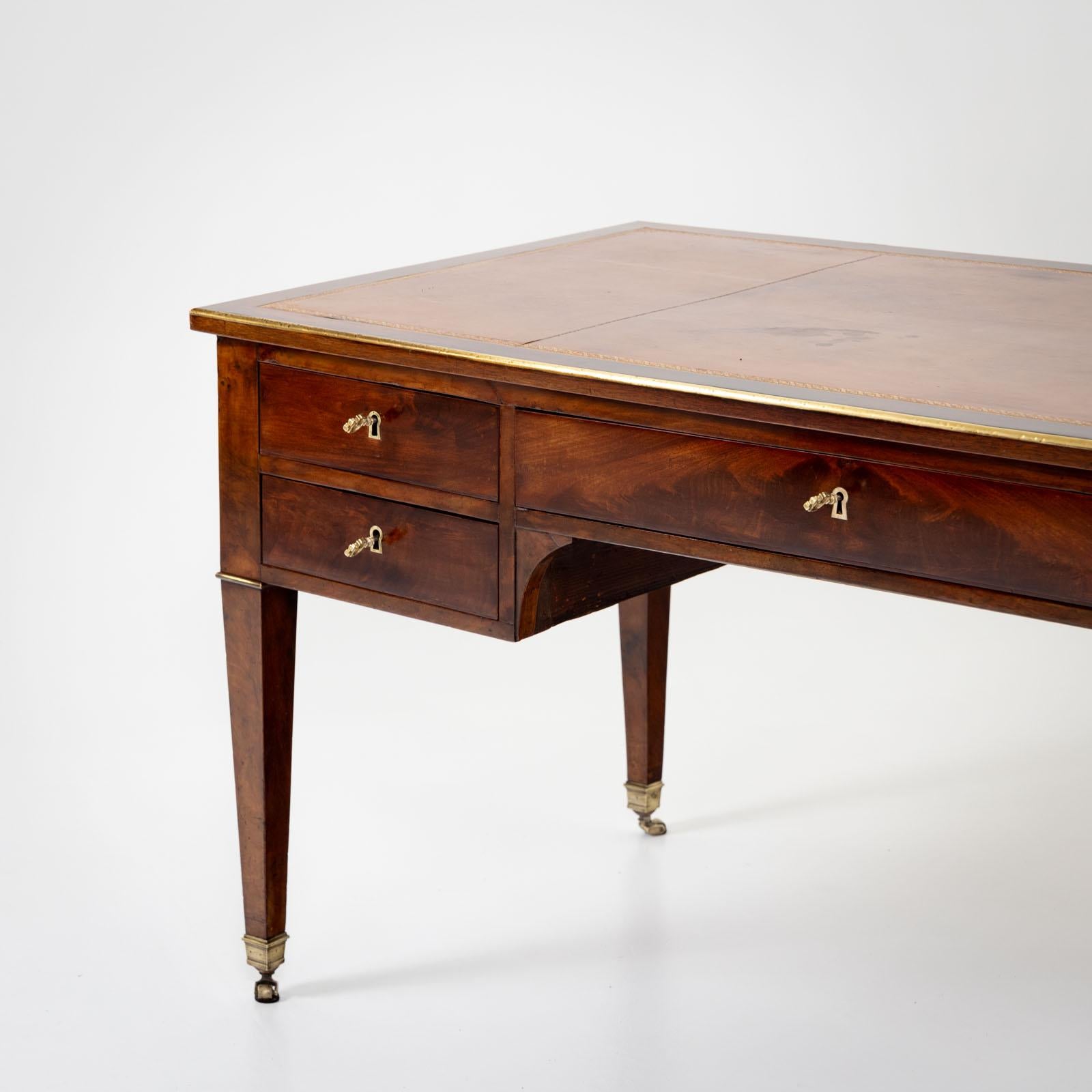 French bureau plat on square tapered legs with brass sabots and small castors. The desk is veneered in mahogany and the writing surface is covered in congac-colored leather. The keyholes are lined with brass and the tabletop is edged with a brass