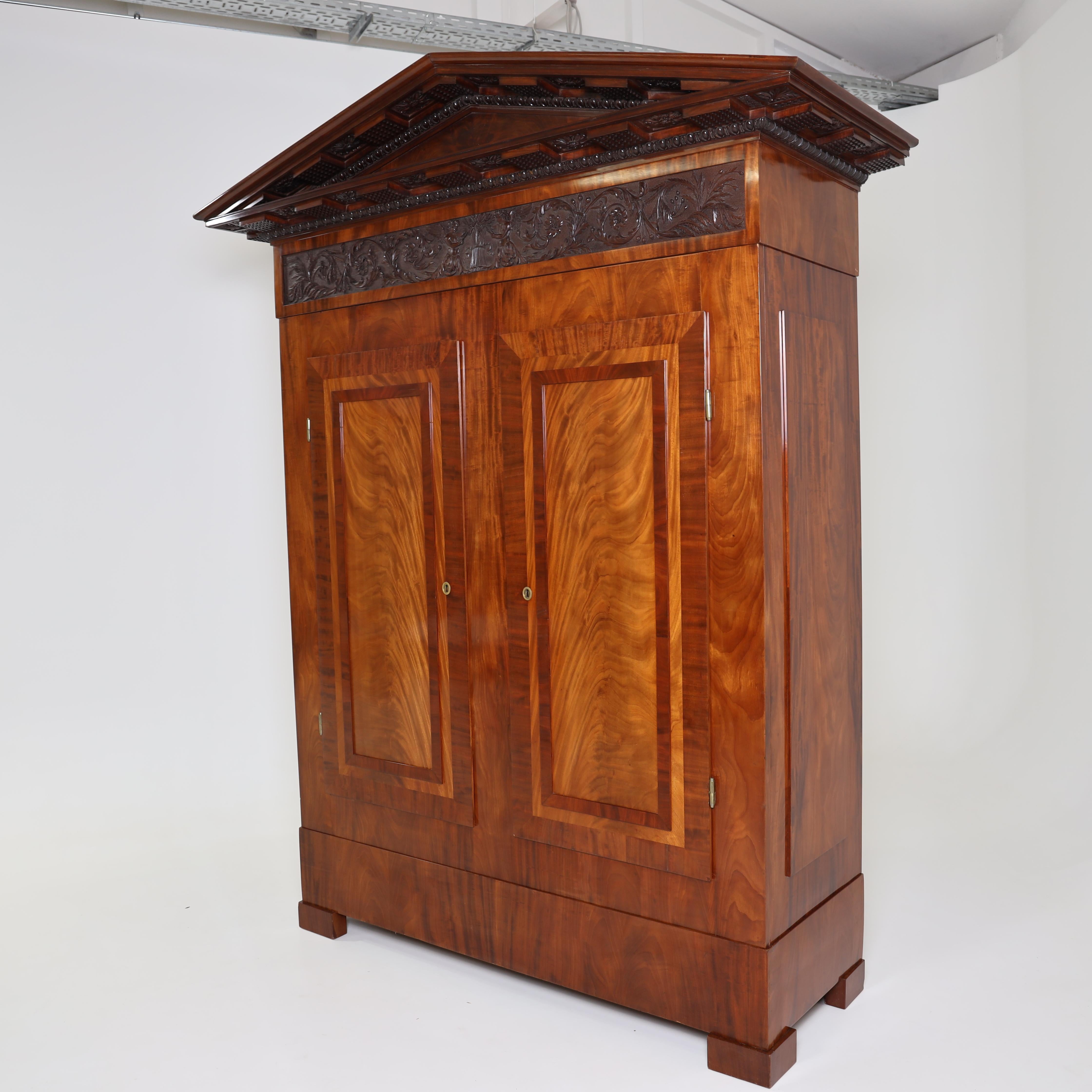 Two-door cabinet with smooth front with panels veneered in mahogany. The smooth plinth rests on low square feet. The temple-like pediment with smooth tympanum field is visually set off with a carved and vine decorated frieze and framed by a