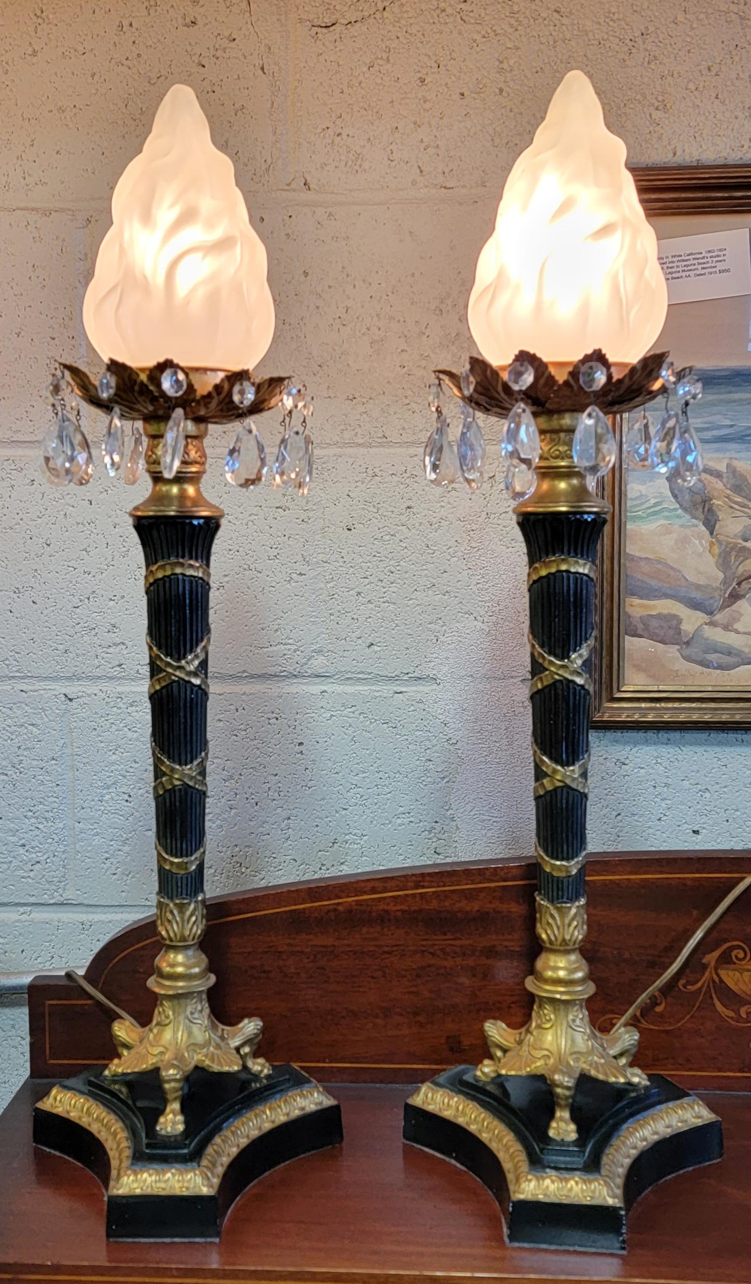 Striking pair of Neoclassical table lamps. Crystal teardrop prisms surround frosted glass flame shades. Draped candlestick style on footed base. Brass plate and painted metal construction. Mid 20th century. In fine working order.