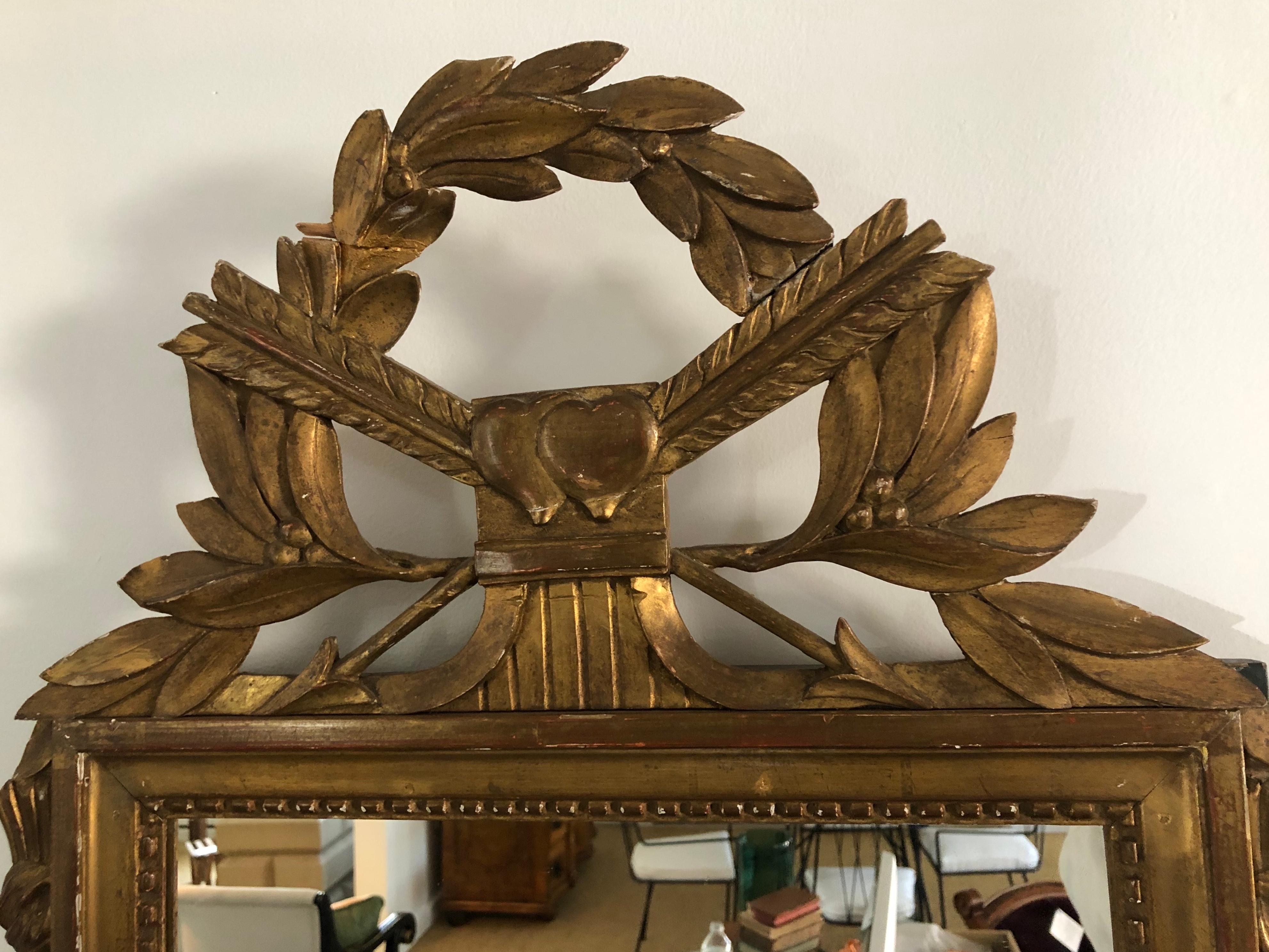 A carved and giltwood neoclassical mirror, the cresting featuring laurel leaves, crossed arrows and 2 overlapping hearts above the rectangular mirror framed with a beaded edge and flanked by a tied tassel on each side, the bottom with green painted