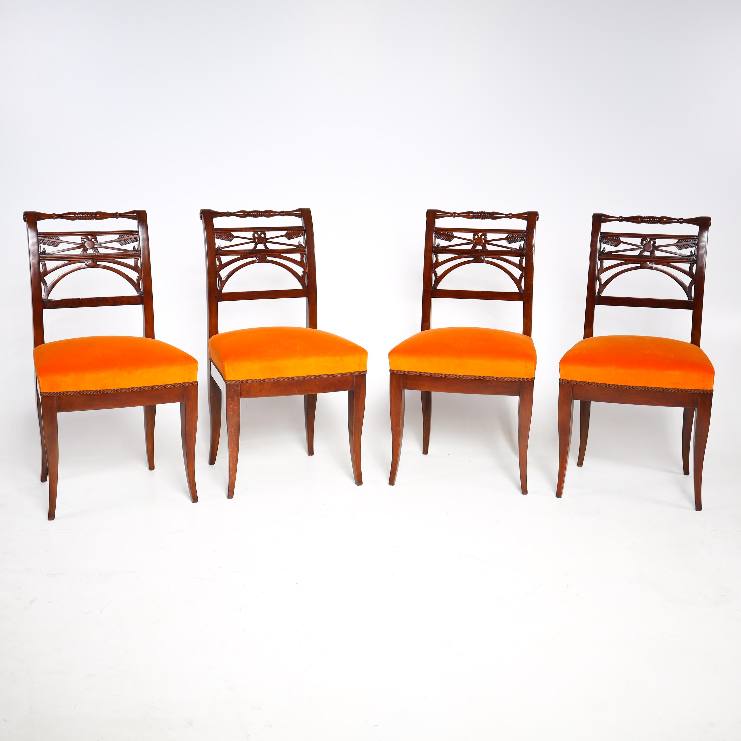 Set of four neoclassical dining room chairs in mahogany, on slightly flared square pointed legs, with straight rails and openwork backrests with bows and arrow decorations. The chairs have been newly covered with an orange fabric.