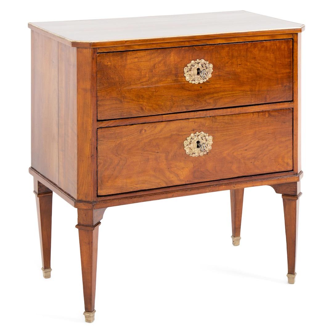 Small neoclassical chest of drawers on square tapered feet with brass sabots. The corpus with beveled corners and two drawers with floral bronze appliqués as escutcheons. A narrow brass strip surrounds the edge of the top plate. Very nice patina