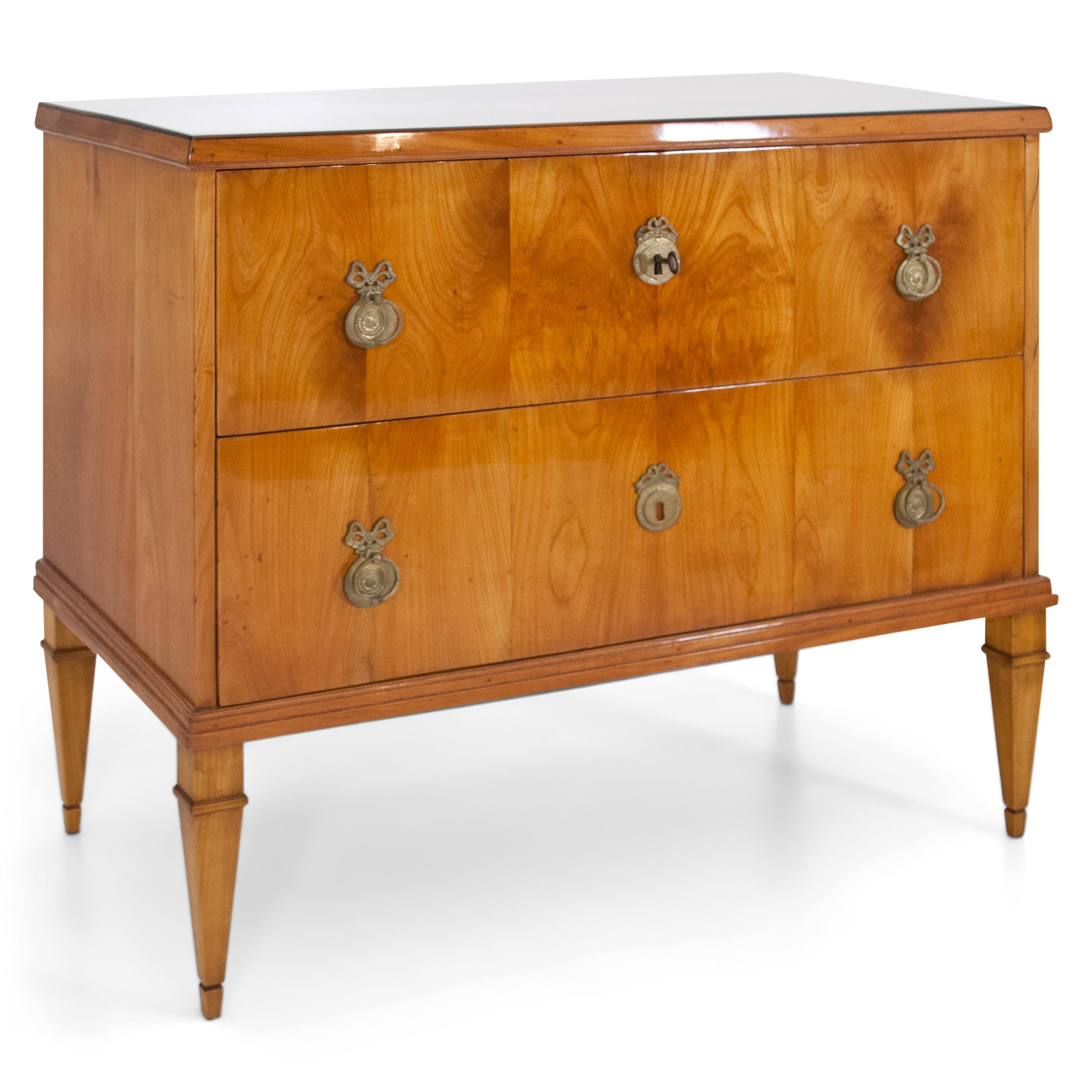 Two-drawered chest of drawers standing on square-pointed feet with profiled lower rail and bronze fittings with pull rings and ribbon decoration. Cherry veneered. The feet are recent. The chest of drawers has been restored and hand polished.
 