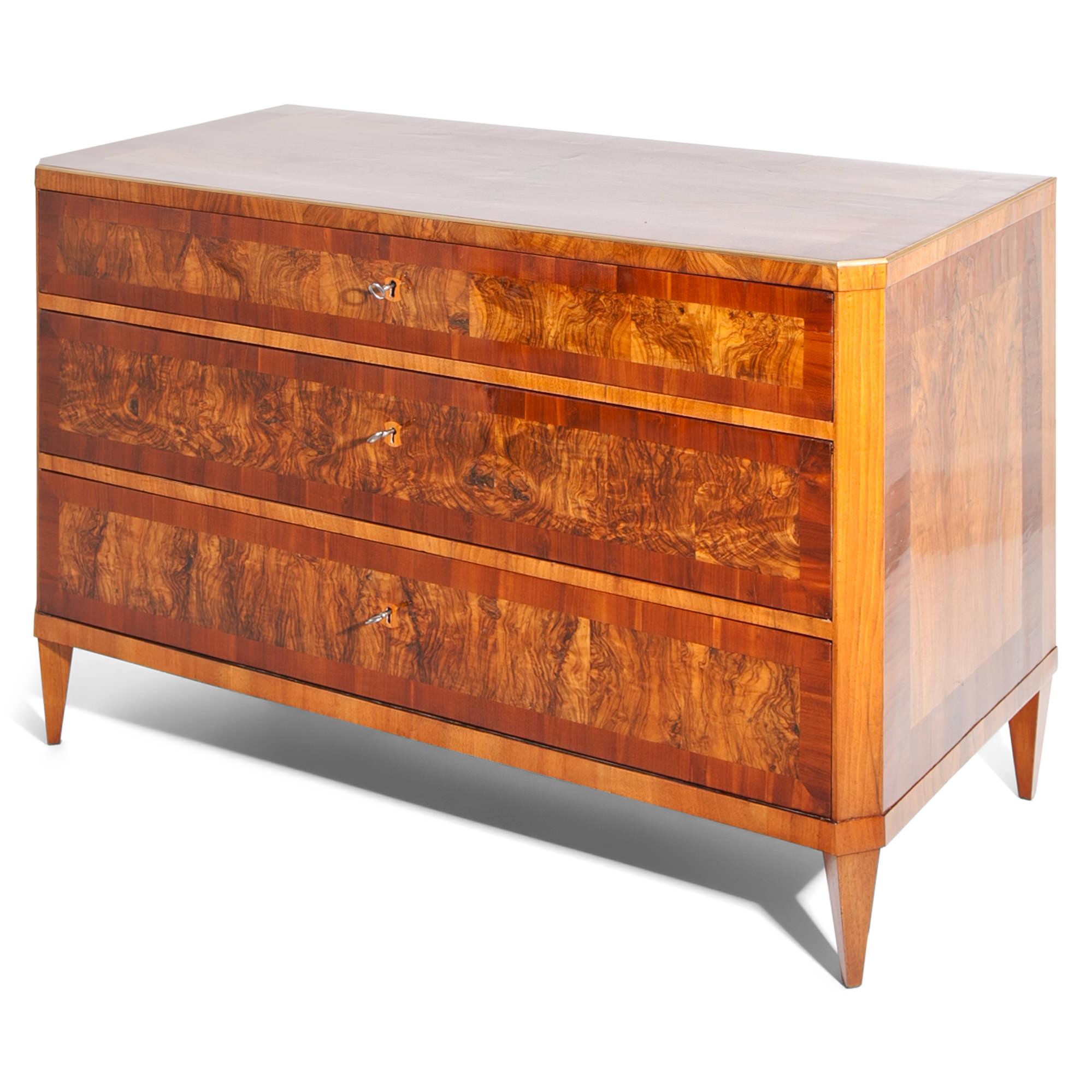 Three-drawered neoclassical chest of drawers with beveled corners, standing on tapered feet. The upper edge is decorated with a brass strip. The front and sides are veneered in burl wood and walnut. The chest was professionally refurbished and