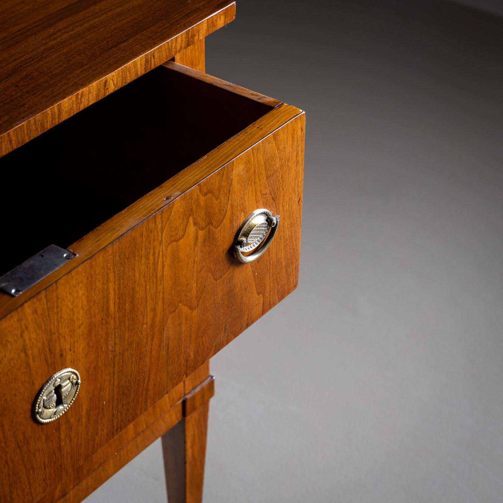 Piccola cassettiera neoclassica con due cassetti e accessori in ottone. L'impiallacciatura è stata lucidata a mano. Le gambe affusolate con sabot in ottone aggiungono un tocco elegante all'estetica minimalista.