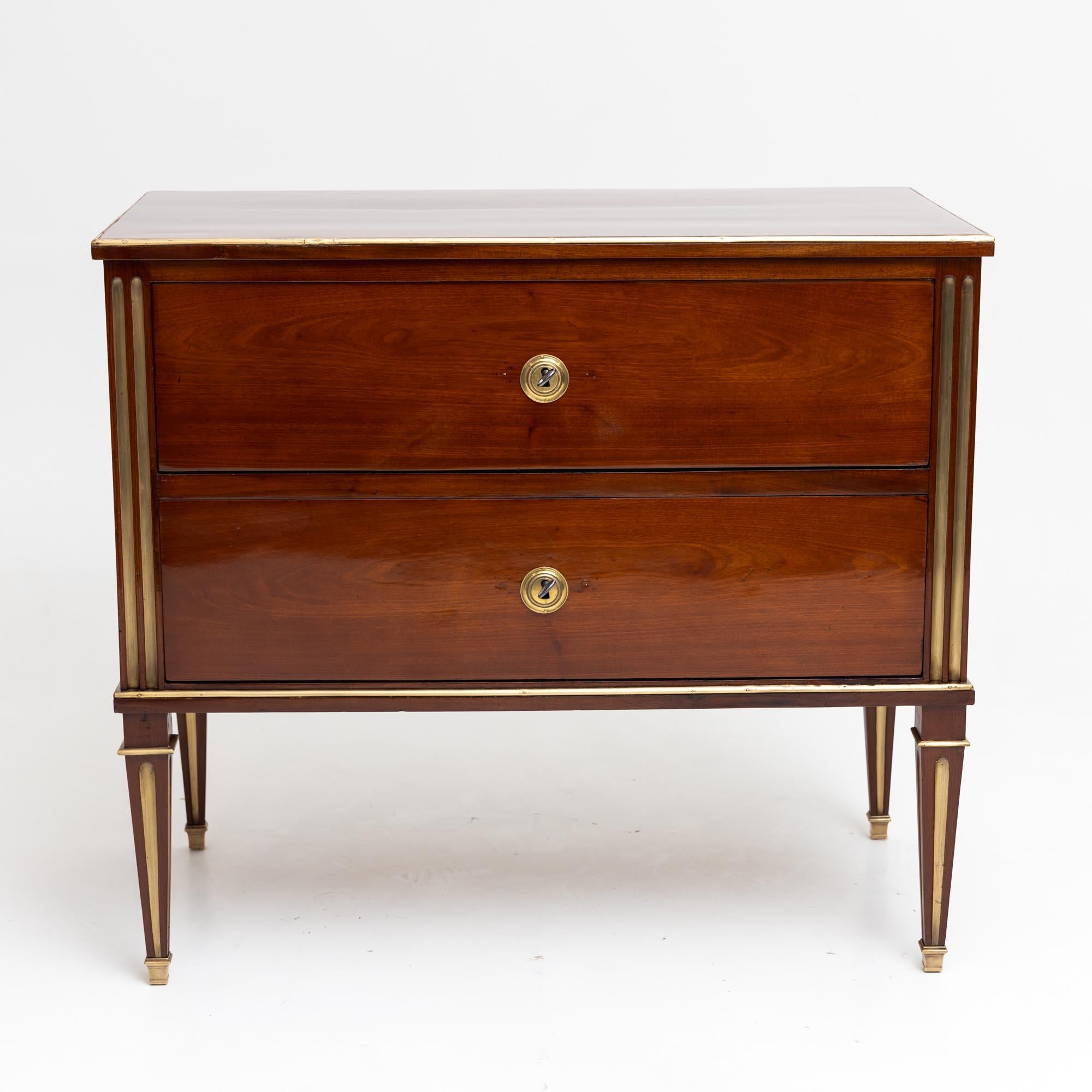 Neoclassical chest of drawers with two drawers and brass profiles. The pilaster strips are fluted and also accentuated in brass. The chest of drawers stands on square pointed feet with brass sabots and is veneered in mahogany. The chest of drawers