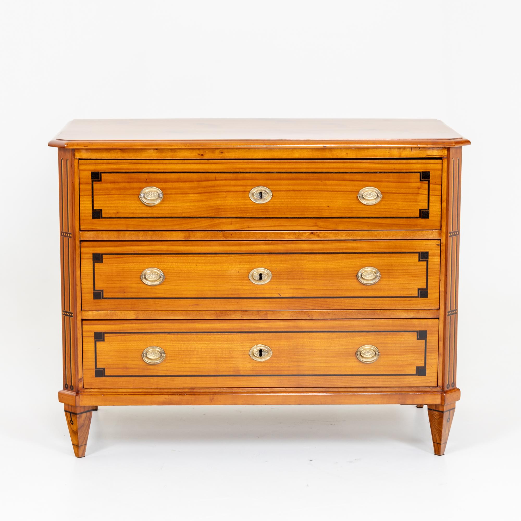Neoclassical chest of drawers on square pointed feet with three drawers and oval bronze fittings. The corpus is decorated with thread inlays on the bevelled corners and the fronts of the drawers. The cherry veneered chest of drawers has been