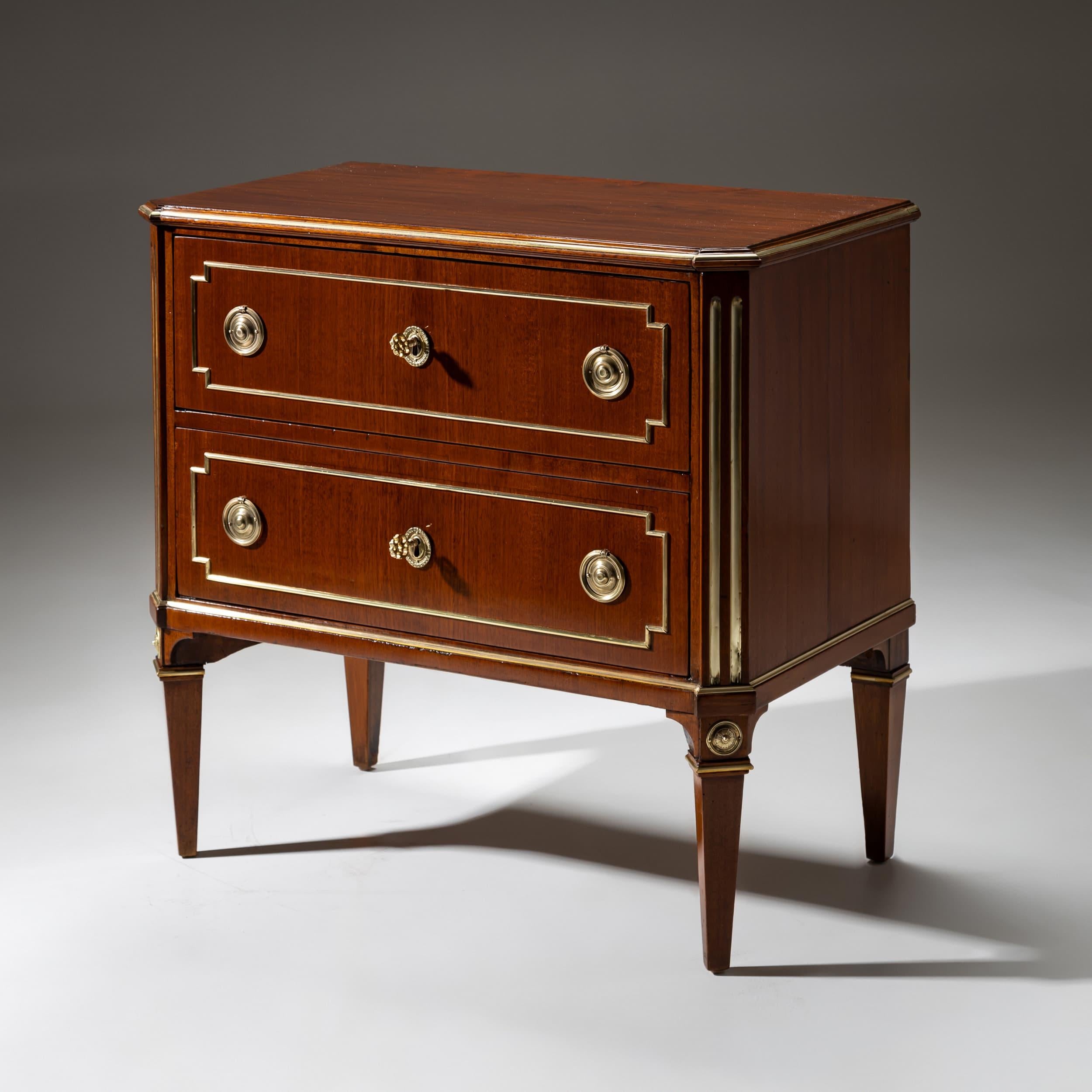 Commodo neoclassico a due cassetti con angoli smussati e gambe affusolate. Gli angoli, le gambe, i frontali dei cassetti e i bordi del corpo sono impreziositi da profili in ottone. Il corpo è impiallacciato in mogano e presenta un design simmetrico