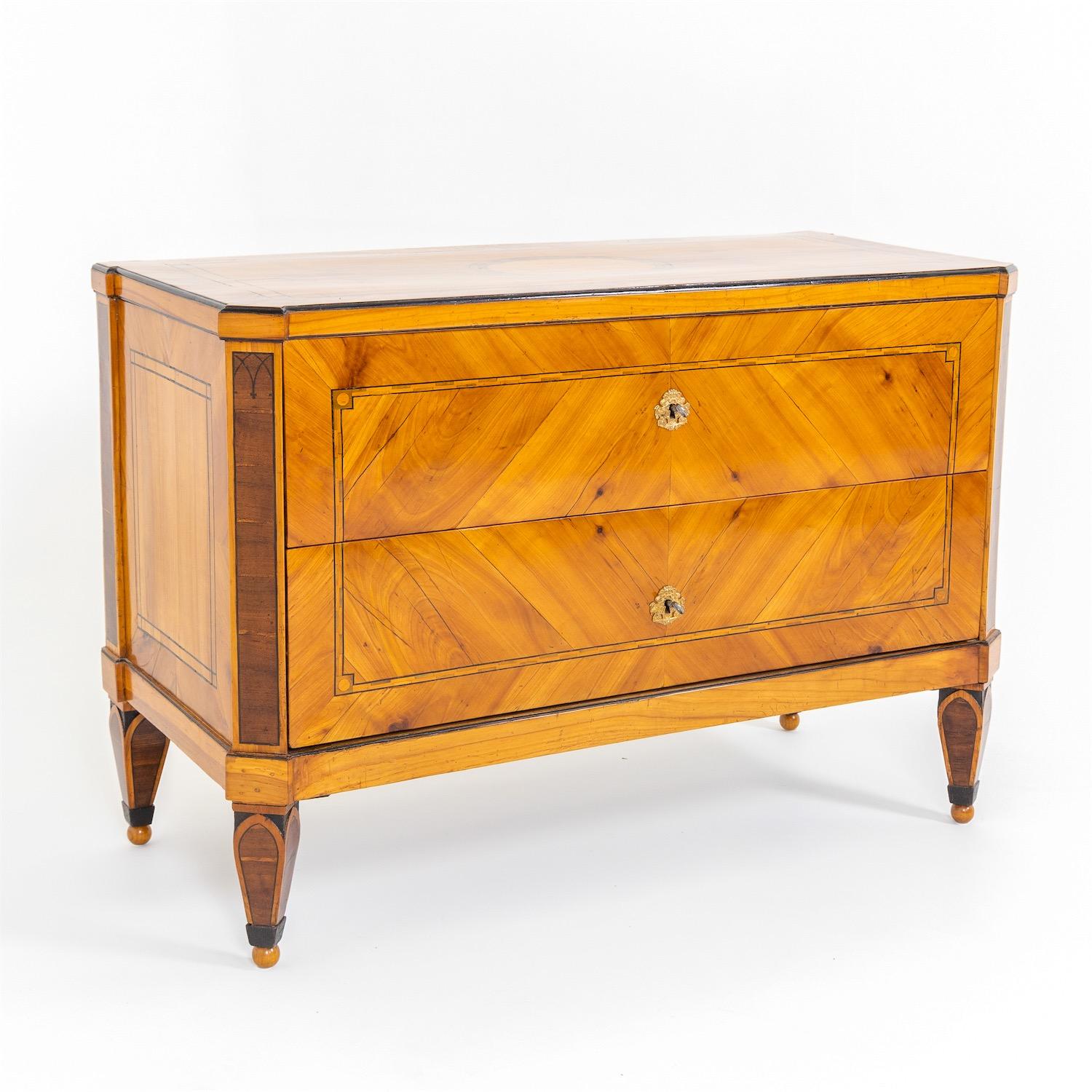 Neoclassical chest of drawers on square pointed legs with accentuated arche shapes and ball feet. The beveled, slightly cranked corners are decorated with additional lancette arches and inlaid in dark stained wood. The two drawers are constructed