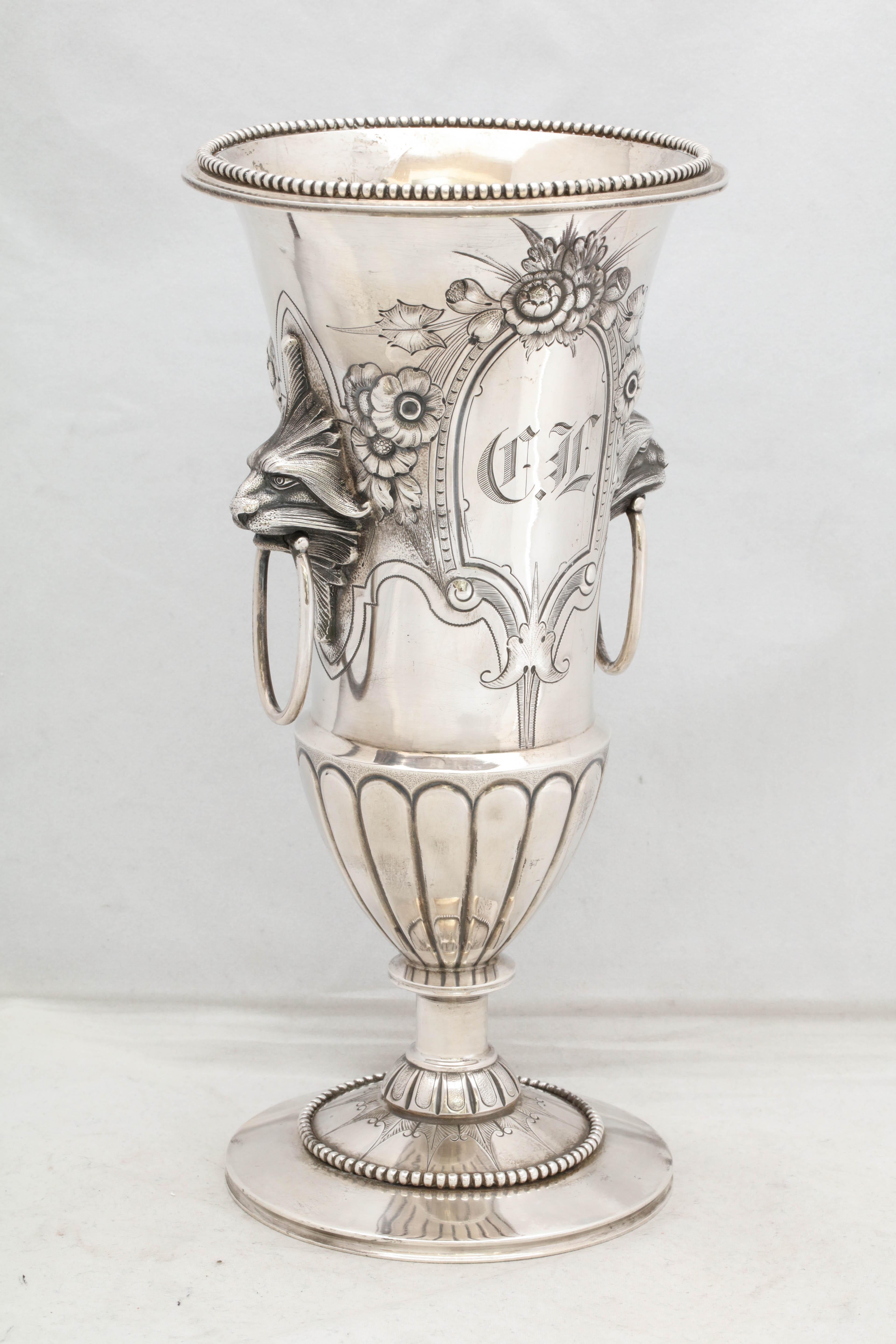 Neoclassical, coin Silver, pedestal - based vase, New York, circa 1875, Wood and Hughes - Makers. On each side of the vase, a ringed handle emanates from a lion's mouth. Vase is chased with flowers and is etched. Rim of vase is decorated with