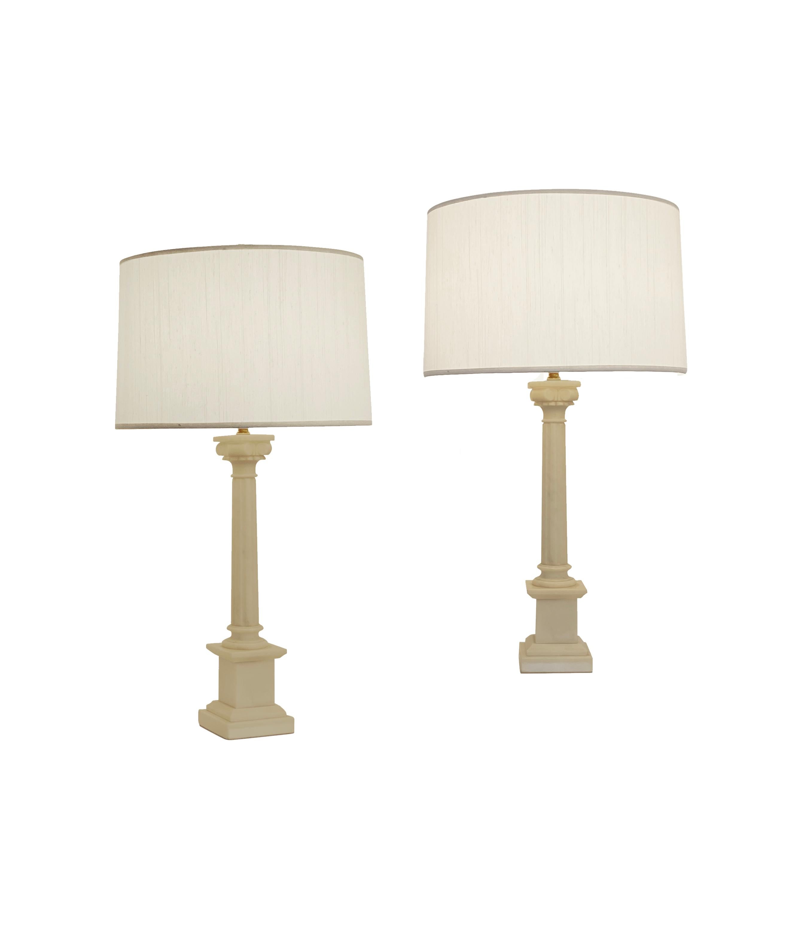 A beautiful pair of alabaster neoclassical Corinthian table lamps with silk ivory lamp shades. Alabaster marble features natural yellowing and some minor chips or scratches throughout given age. Shades are new and lamps also feature new brass