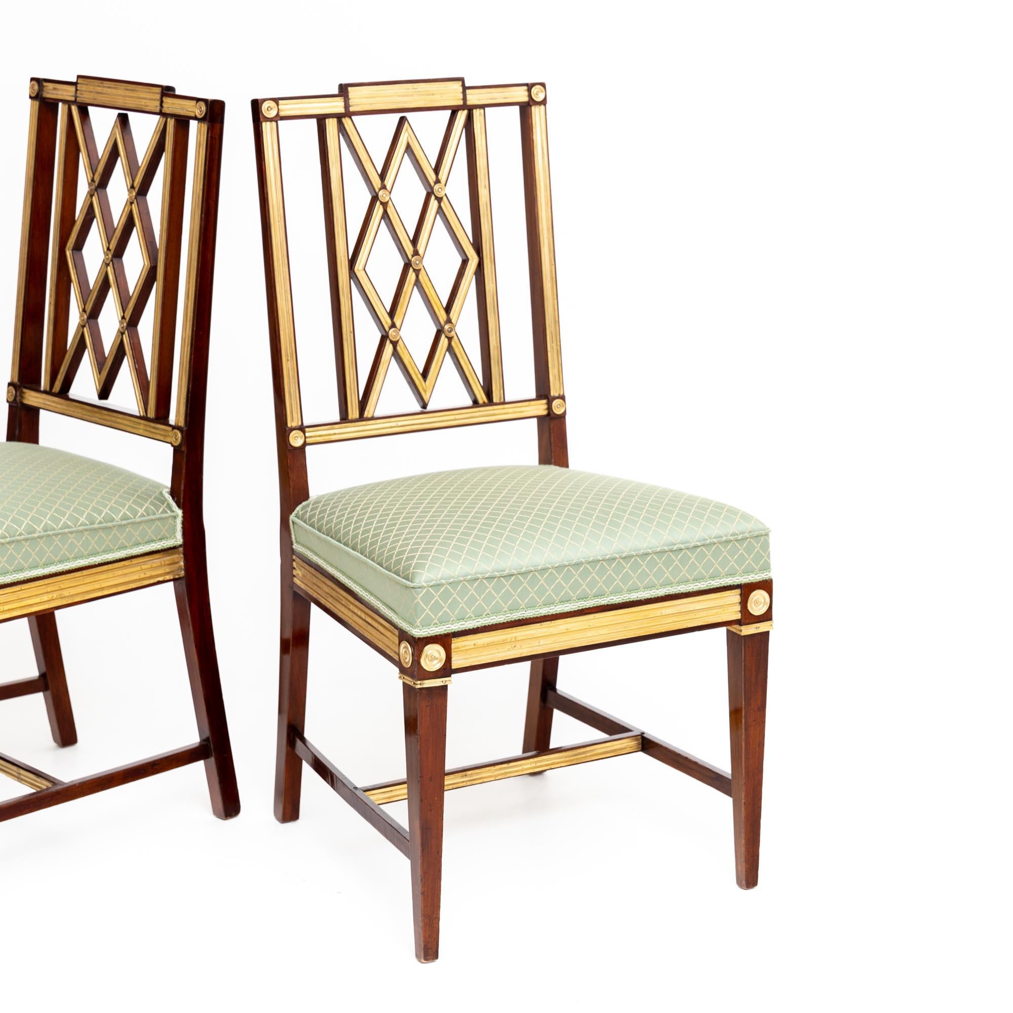 Set of three neoclassical dining chairs made of mahogany with brass decoration and diamond shaped backrest. The chairs stand on square legs with H-shaped bracing. The seats are upholstered and covered with a high-quality light green fabric with a
