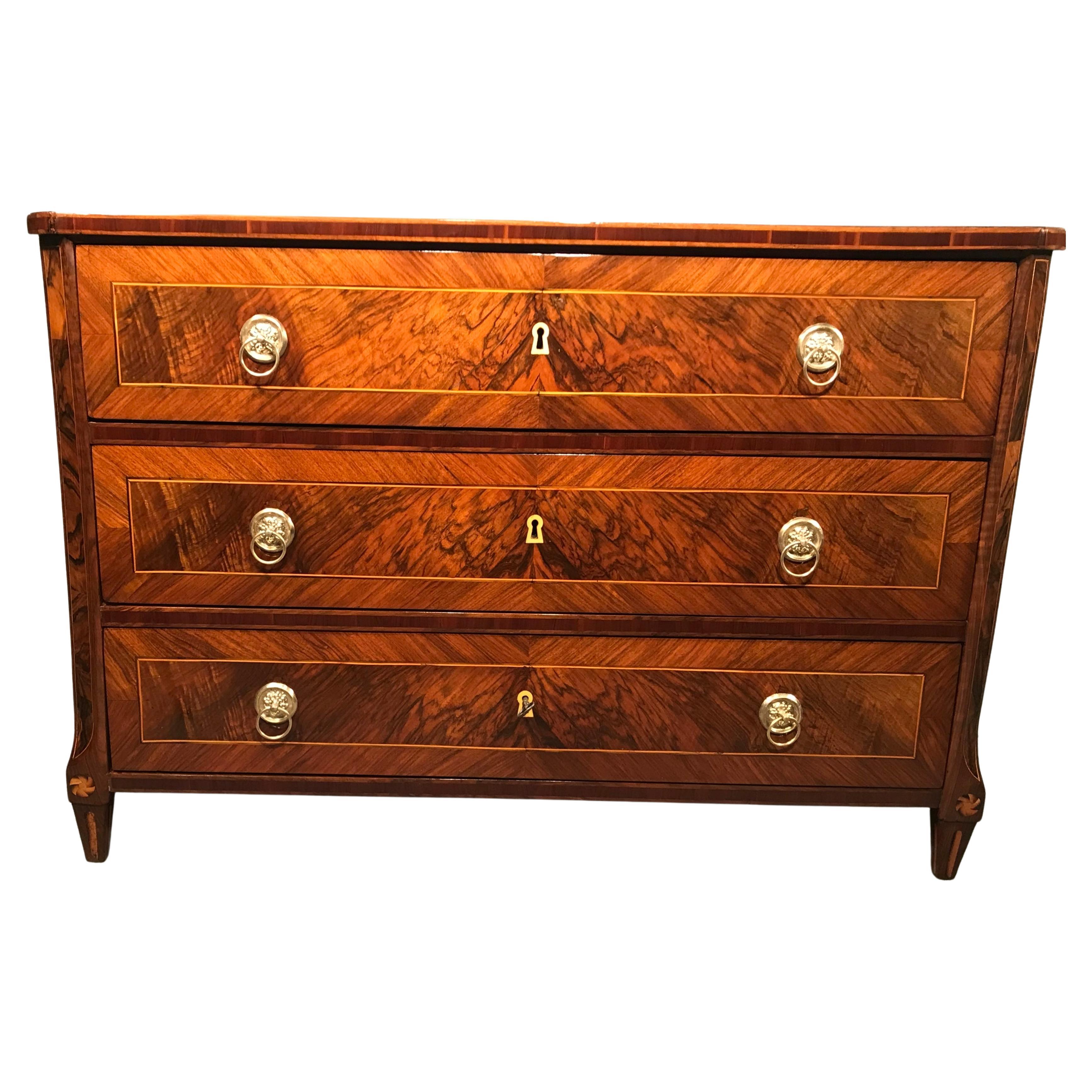 This elegant Neoclassical chest of drawers comes from Germany and dates back to around 1780-1800. 
It stands out for its beautiful variations of walnut veneer grains. The top of the three drawer commode features a mirrored walnut root veneer. The
